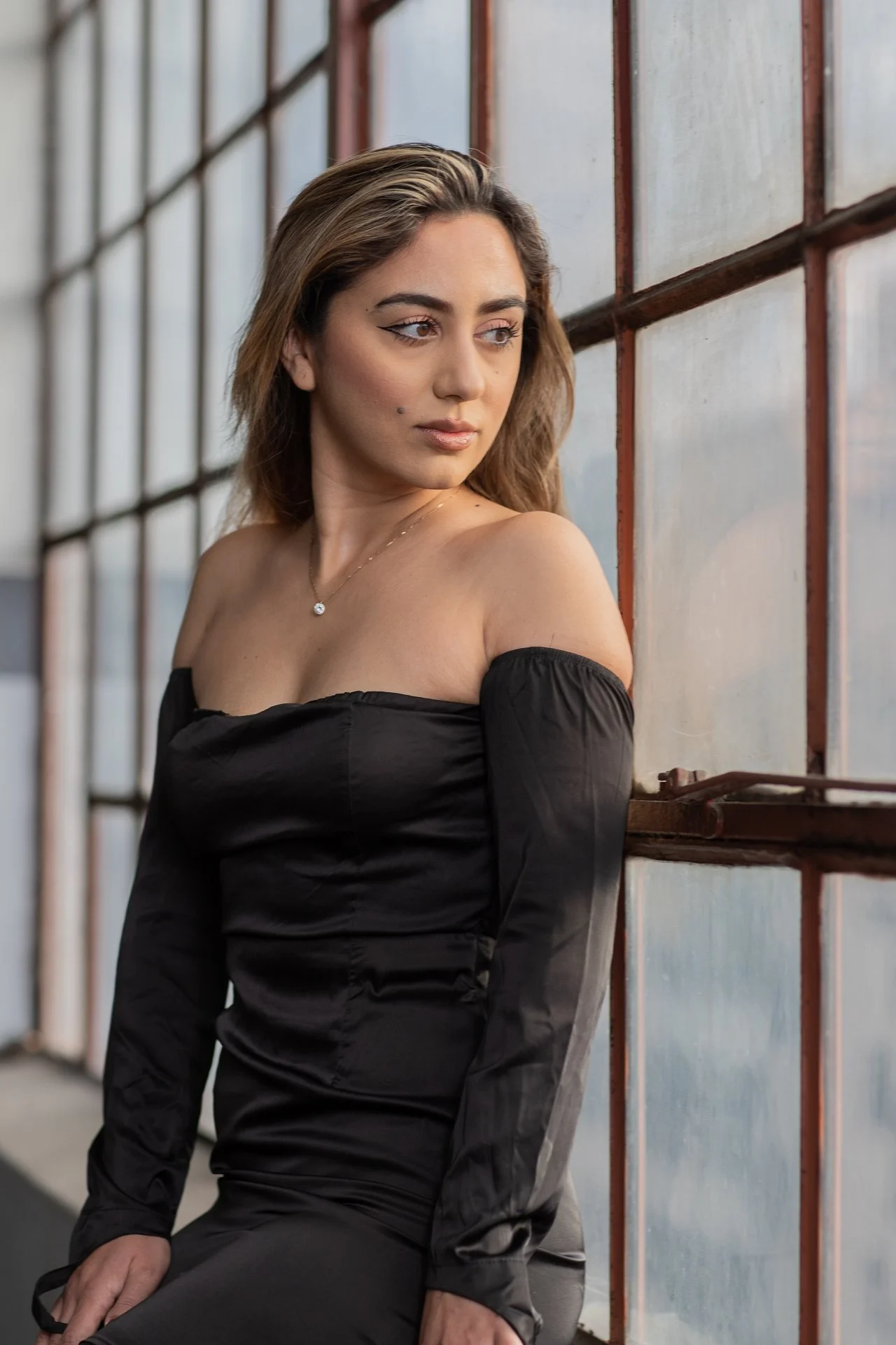 A young woman with wavy brown hair and a small beauty mark on her face, wearing a black off-shoulder top and a delicate necklace, looking thoughtfully to her right while leaning against a window with metal framing.