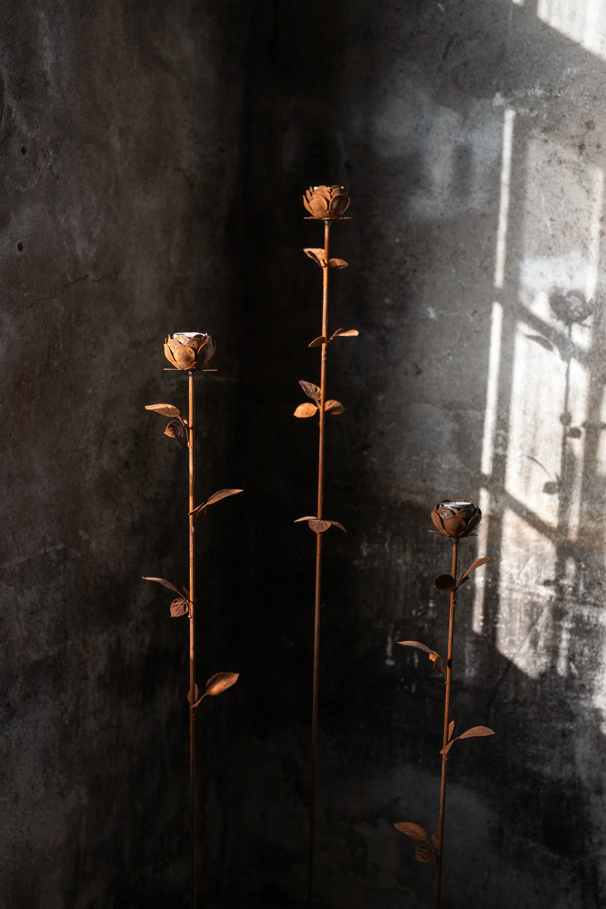 Three tall, rust-colored metal flower sculptures with intricately designed blooms, set against a dark textured wall with light and shadow patterns.