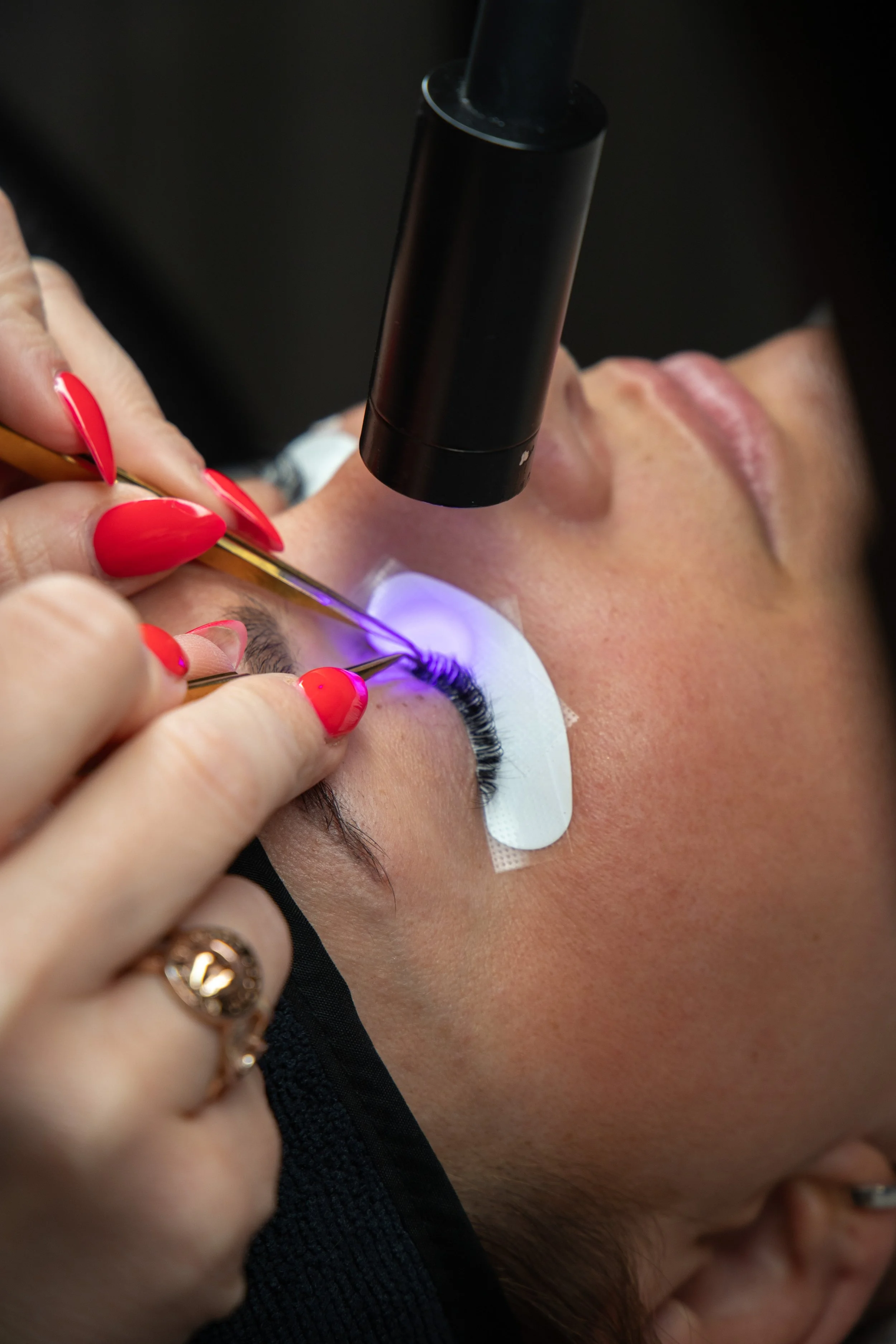 Close-up of a person receiving a purple LED eyelash extension procedure, with technician applying it while the person lounges with eyes closed, protective eye pads, and a magnifying light.
