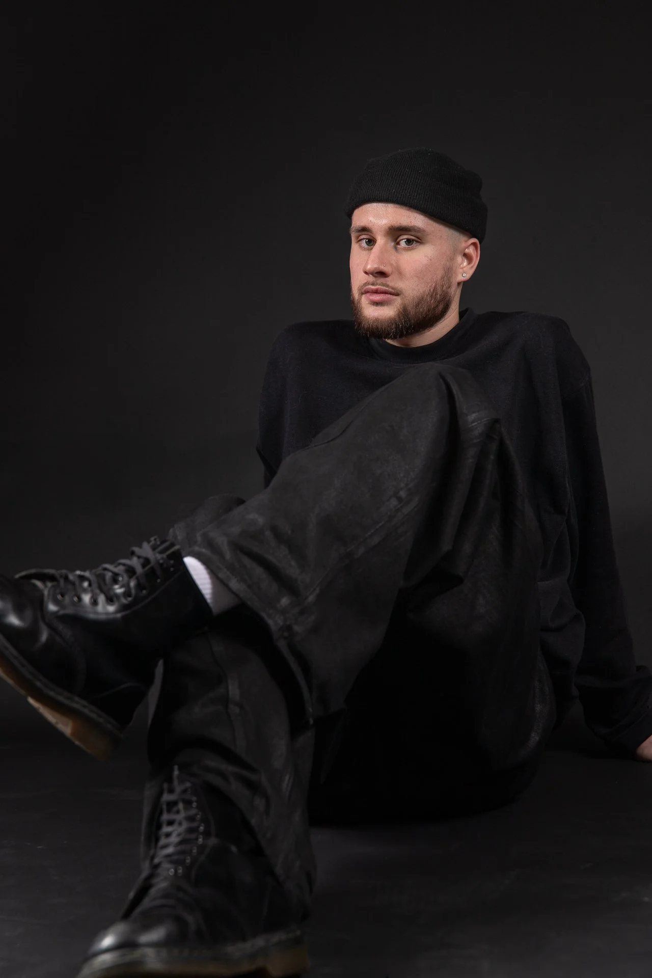 A young man with a beard and light skin sitting down on a dark background, wearing a black beanie, black oversized coat, black pants, and black boots, with his legs extended forward.