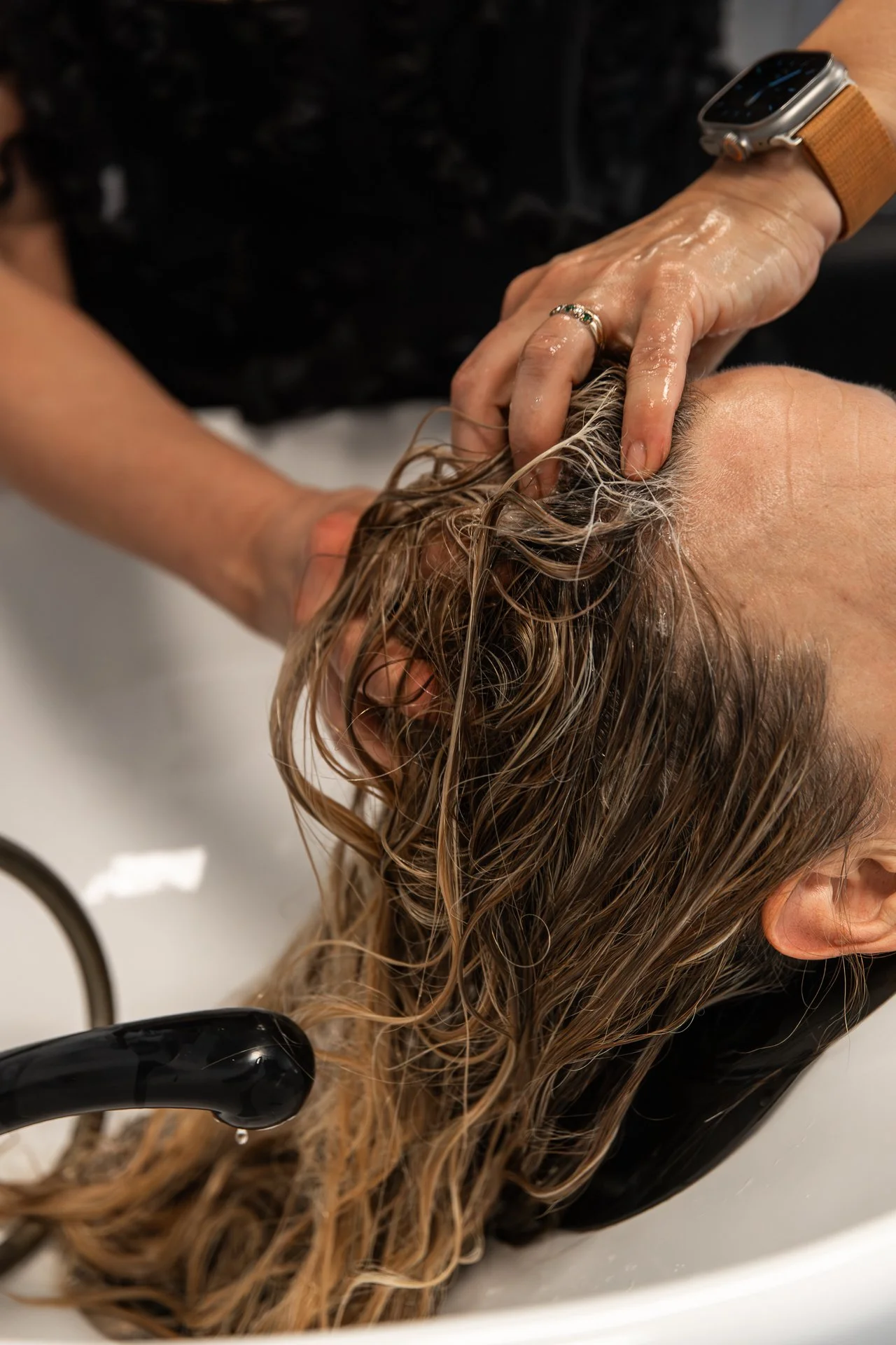 A hairdresser washing a person's wet, curly hair in a salon sink with a black faucet.
