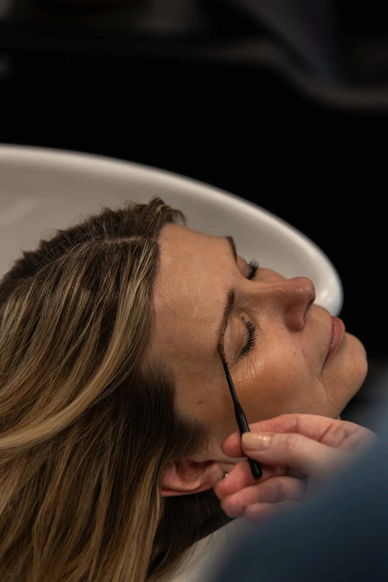 Close-up of a woman receiving an eyelash extension application while lying down with eyes closed. A technician applies the lashes with a fine tool. The woman has long, light brown hair and is resting on a white surface.