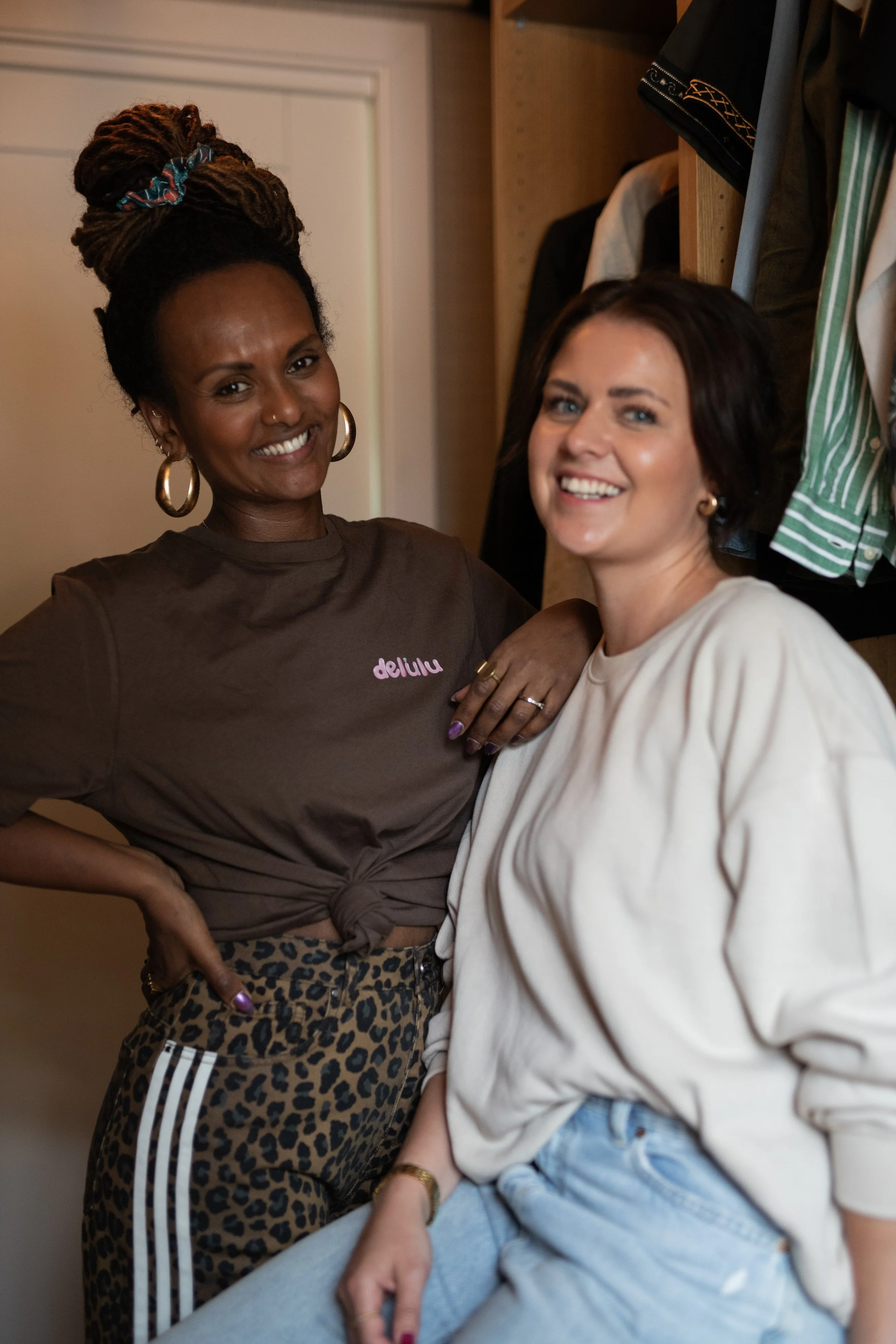 Two women smiling and posing together in a room with clothes hanging behind them.