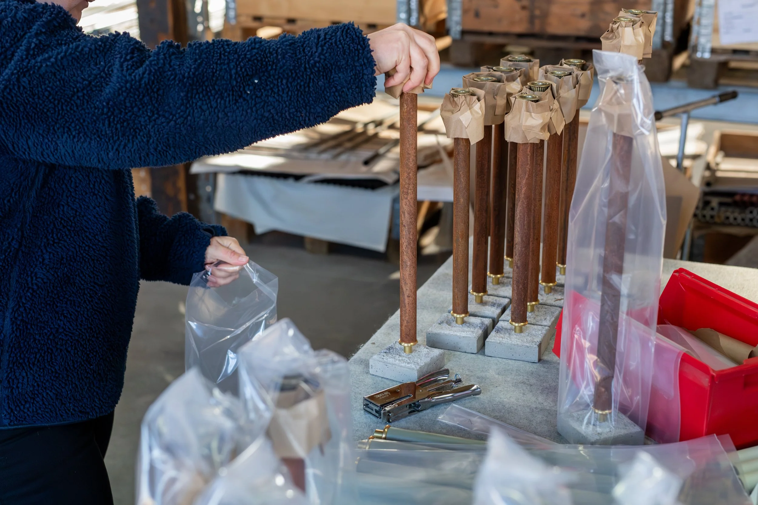 A person working in a workshop or manufacturing setting, assembling copper and concrete pipe structures, with tools and packaging materials on the table.