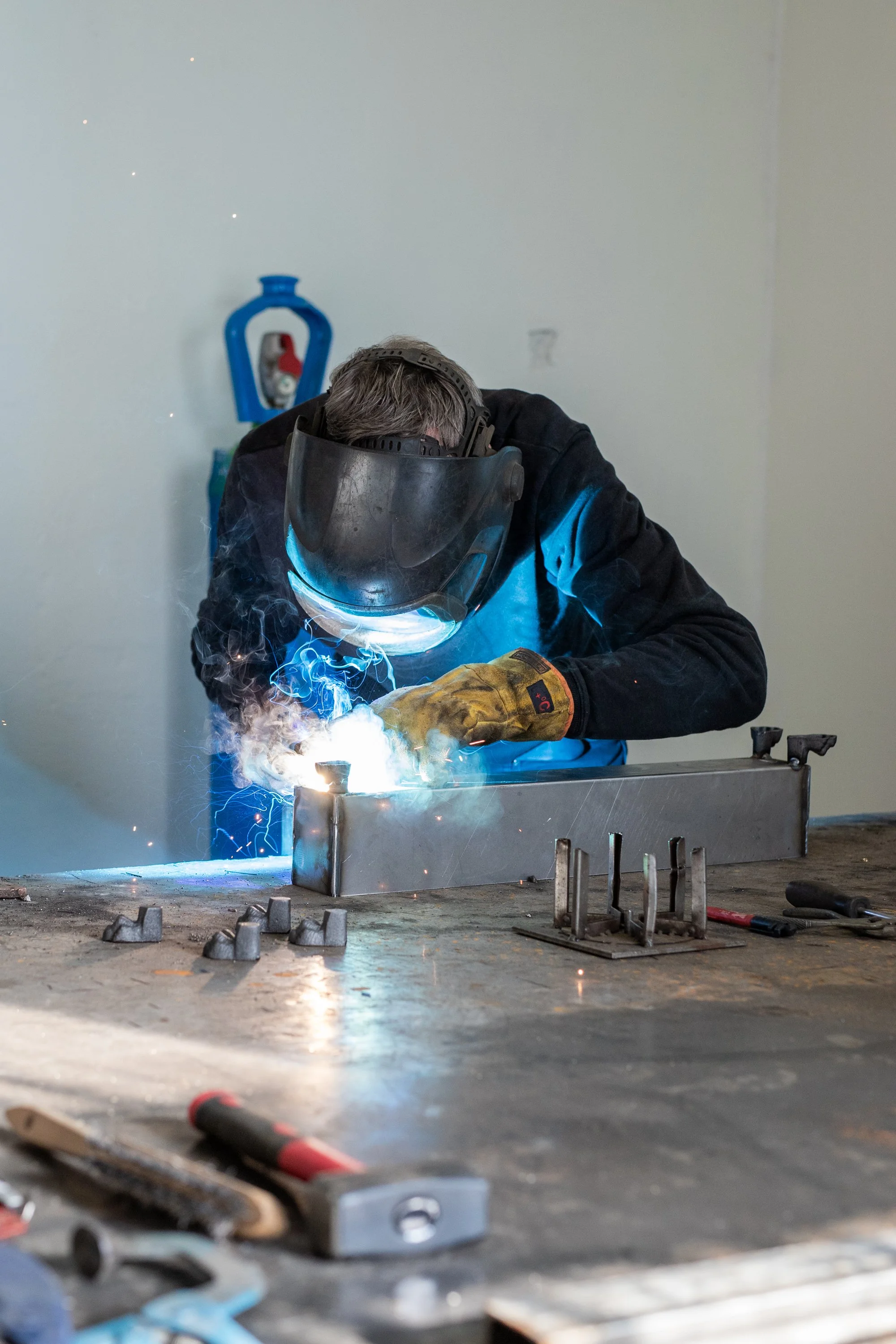 A person welding metal on a workbench using a welding helmet and gloves, with sparks and bright light from the welding process.