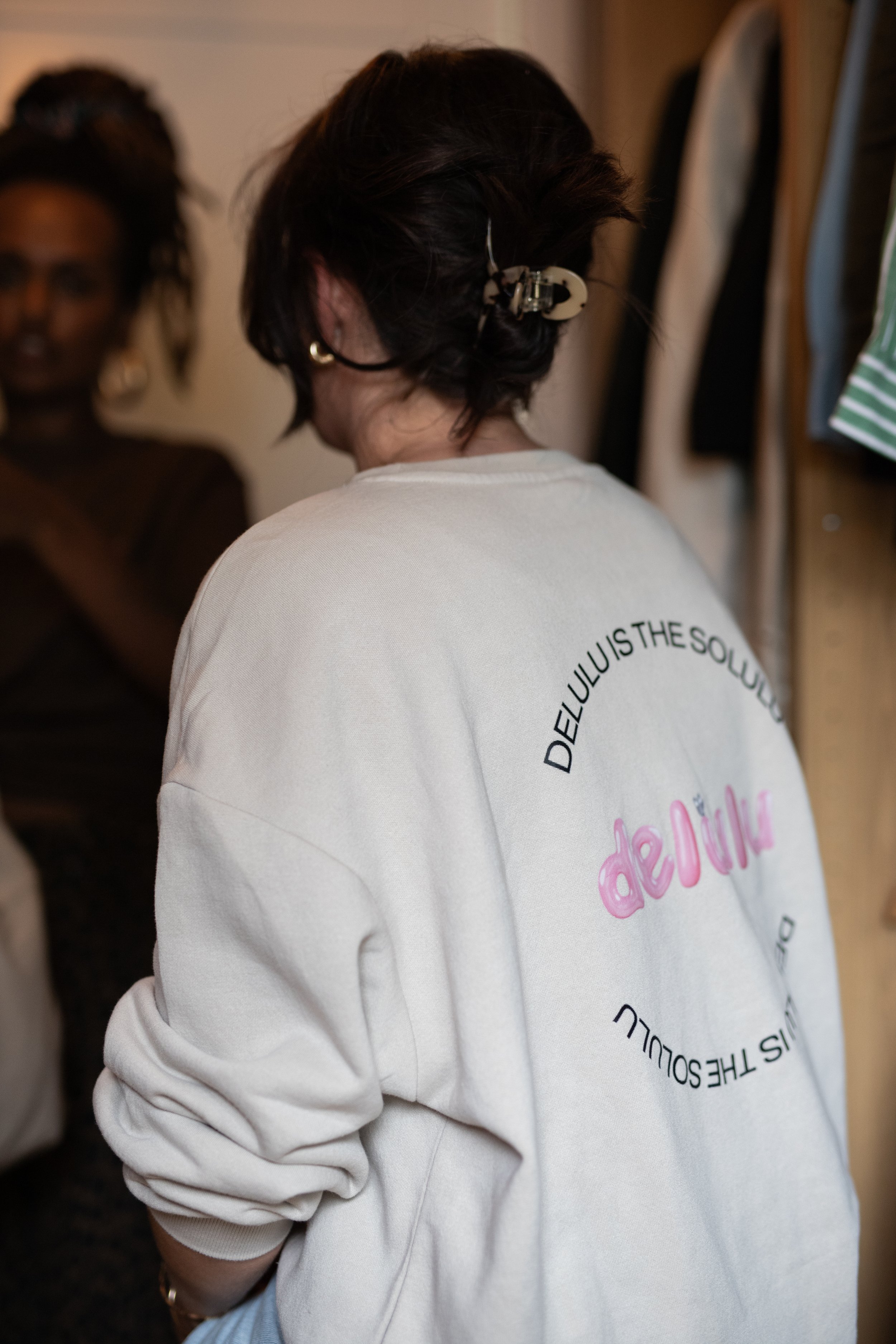 Back of a woman with a short dark hairstyle secured with a hair clip, wearing a white sweatshirt with pink and black text, in a room with another woman in the background.