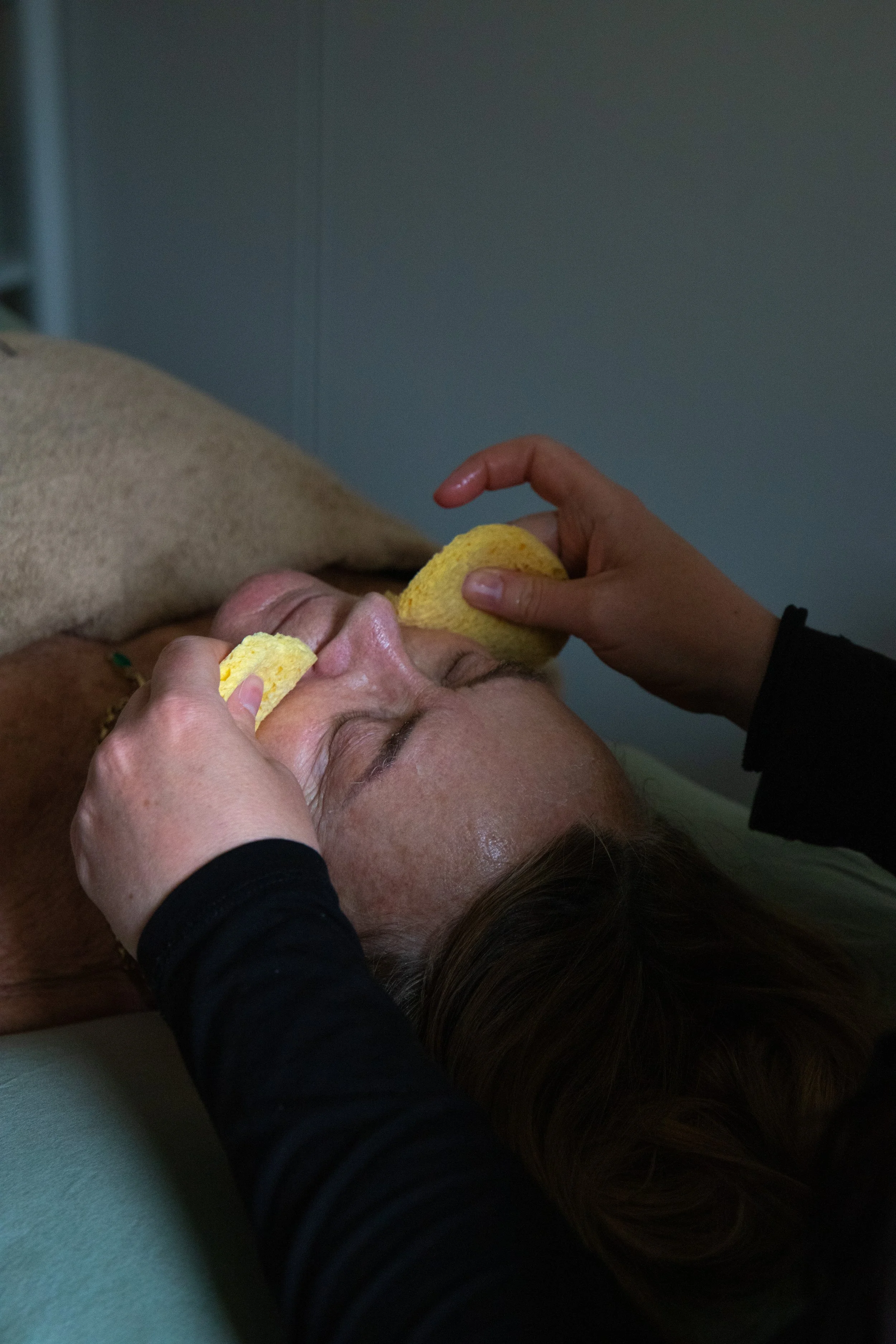 A person receiving a facial treatment while lying down, with another individual applying a product with a yellow sponge to their face.