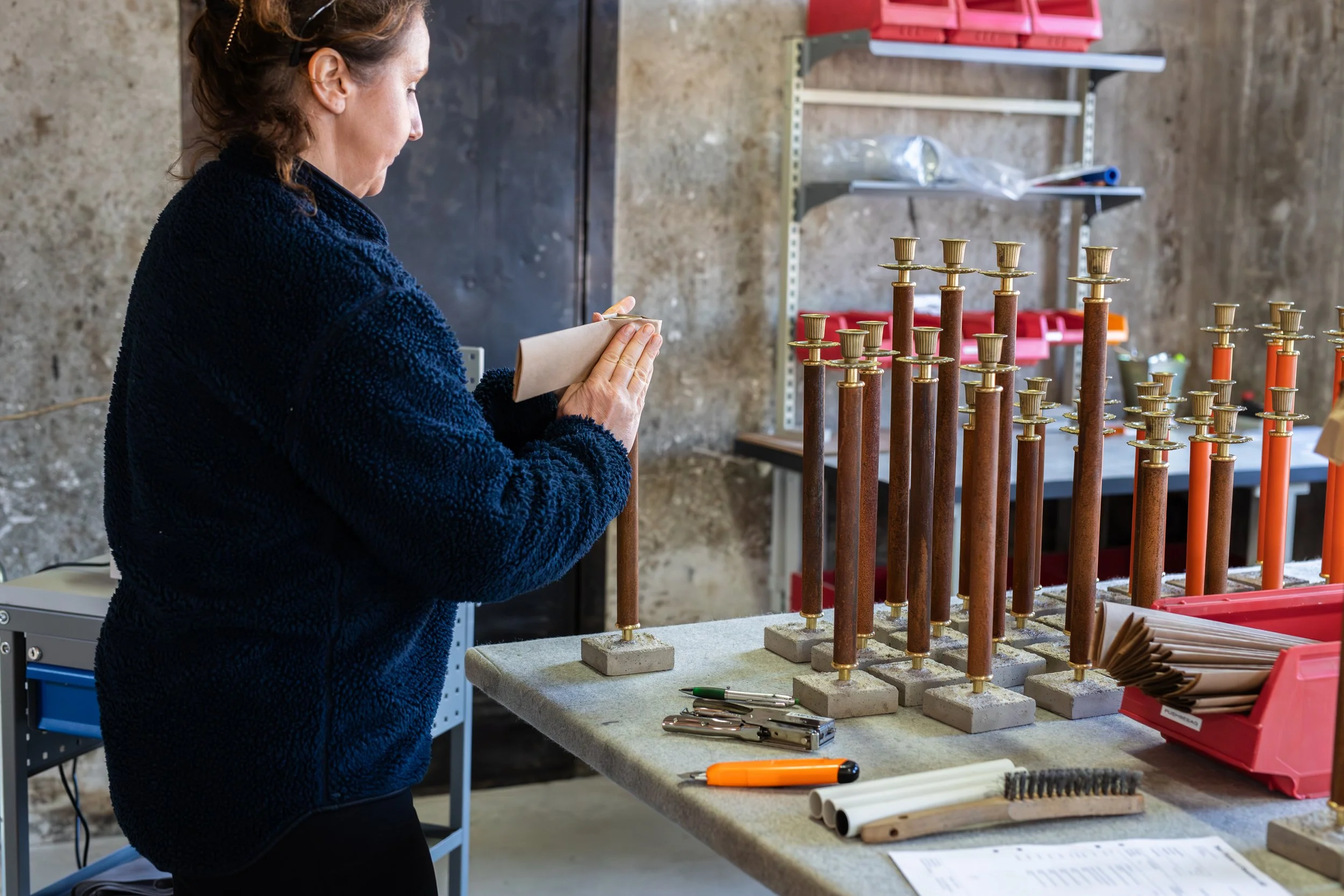 A woman in a dark blue fleece jacket inspects a tall candle with a scope for a candle holder in a workshop. The workspace has a concrete wall and shelves filled with various supplies, with multiple candle holders of different heights on a workbench.