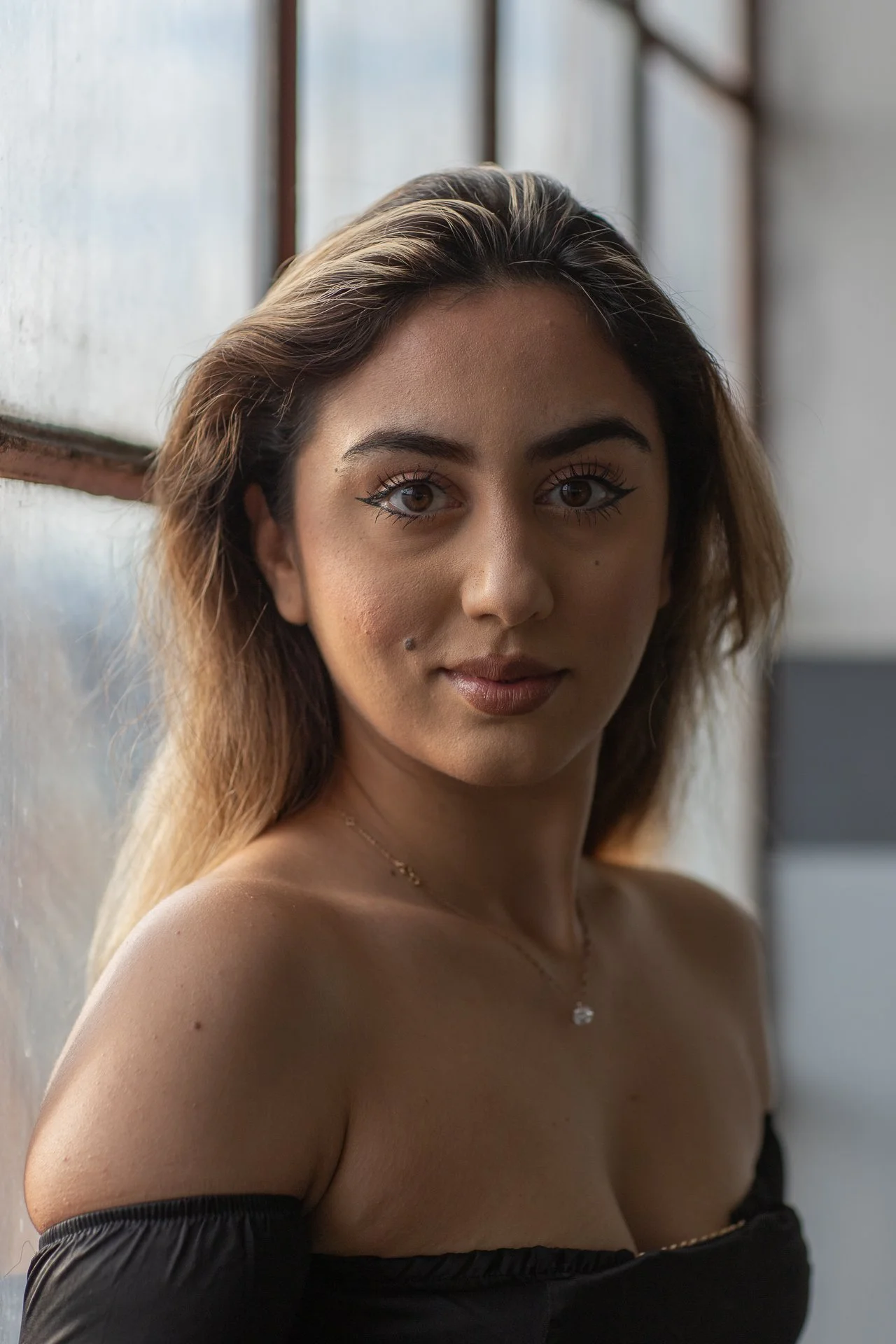 Close-up portrait of a young woman with brown hair, wearing a black off-shoulder top and a delicate necklace. She is looking directly at the camera with a neutral expression, standing near a window with natural light illuminating her face.