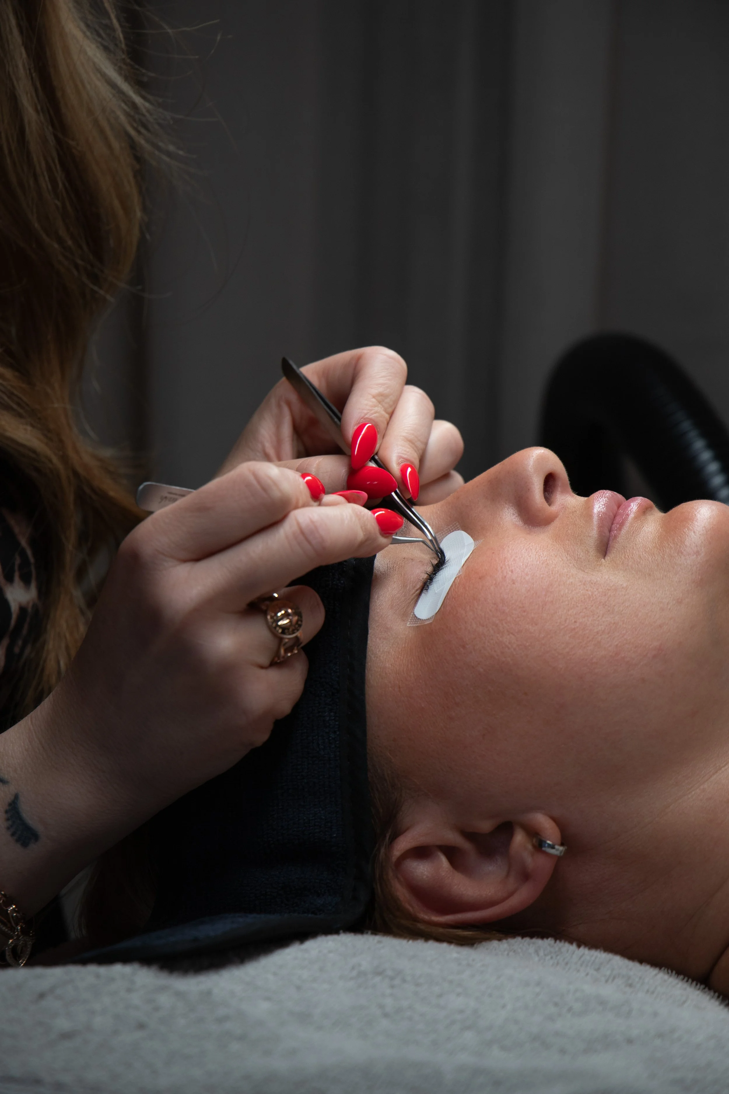 A woman receiving eyelash extension treatment, lying down with her eyes closed while a technician applies eyelashes.