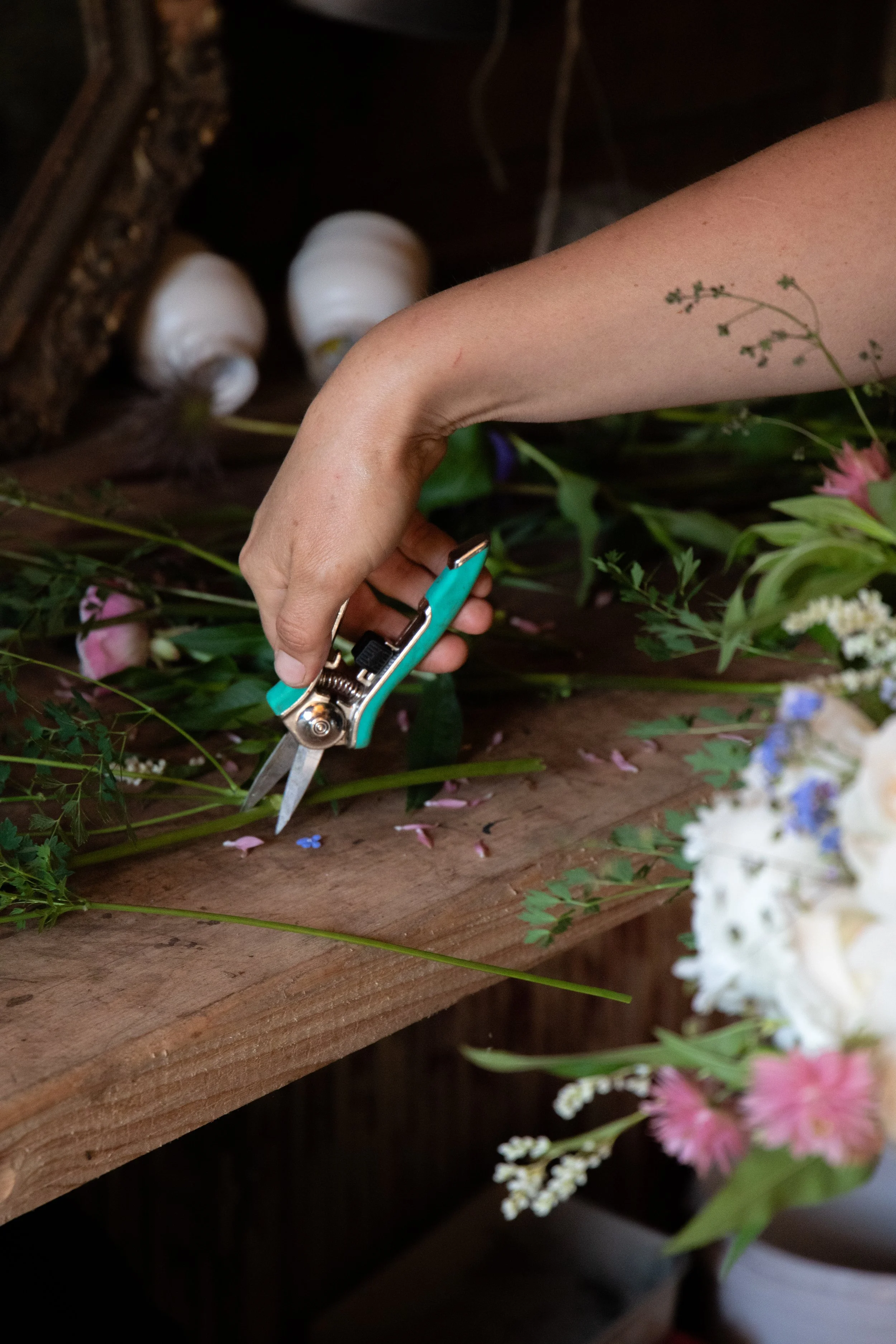 A person's hand is using floral shears to cut flowers on a wooden work surface, with various flowers and foliage around.