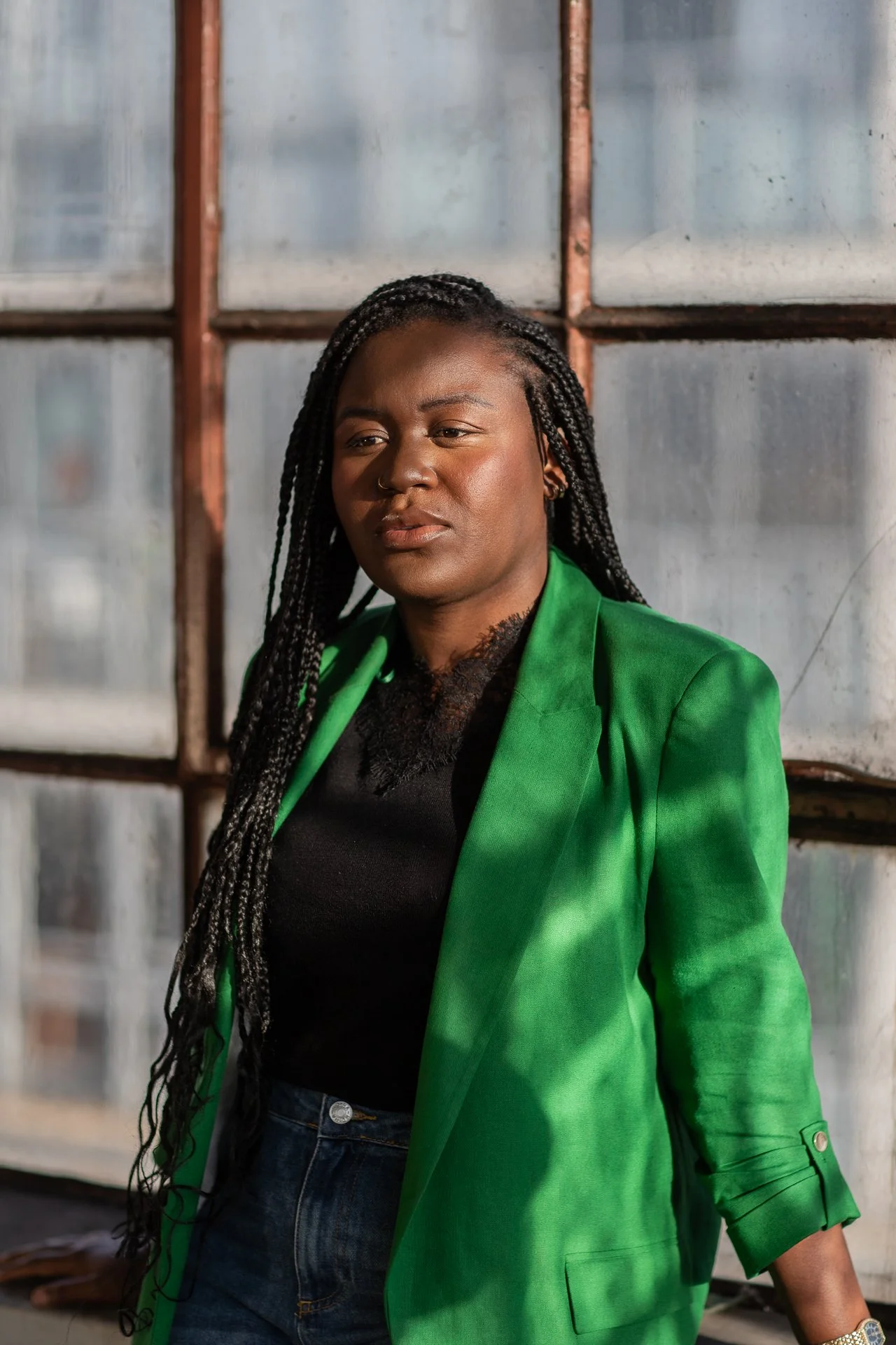 Portrait of a woman with braided hair wearing a green blazer and black top, standing in front of a metal window frame.