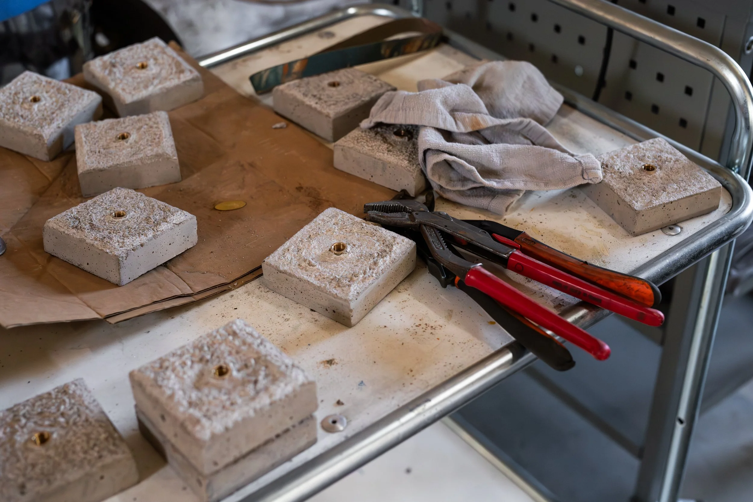 A metal work table with several square concrete blocks, each with a brass fitting in the center, some tools, and cloth rags on it.