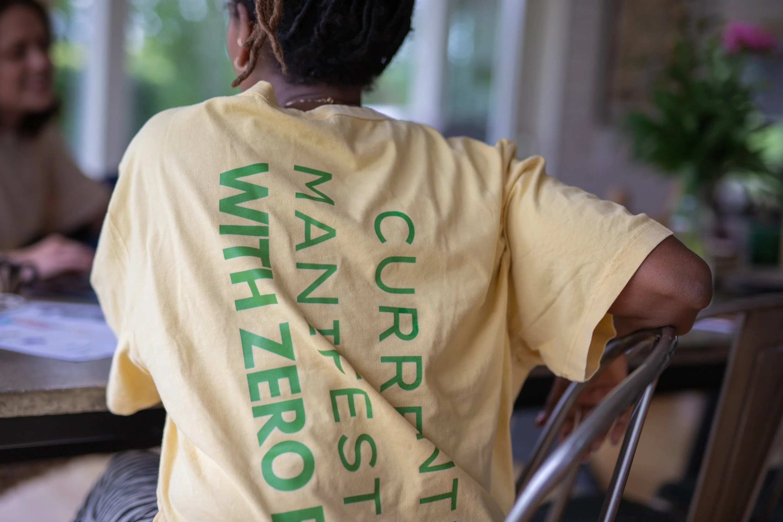 Person wearing a yellow T-shirt with green text that reads "CURESTV CAN'T BE STOPPED", sitting at a table and engaging in conversation with another person.