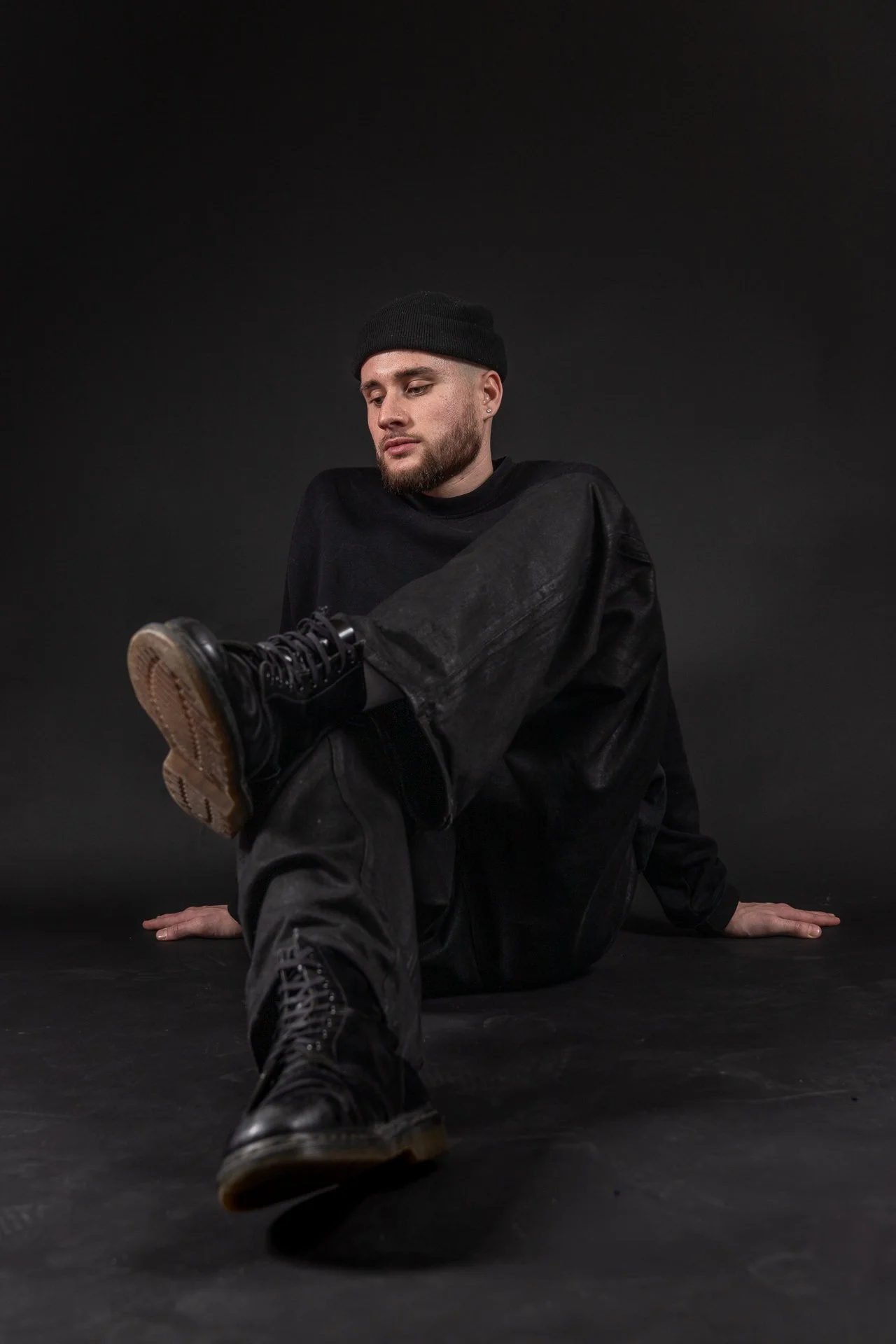 A man wearing black clothing and a black beanie sitting on the floor against a black background, stretching or in a dance pose, with one leg extended and the foot pointed toward the camera.