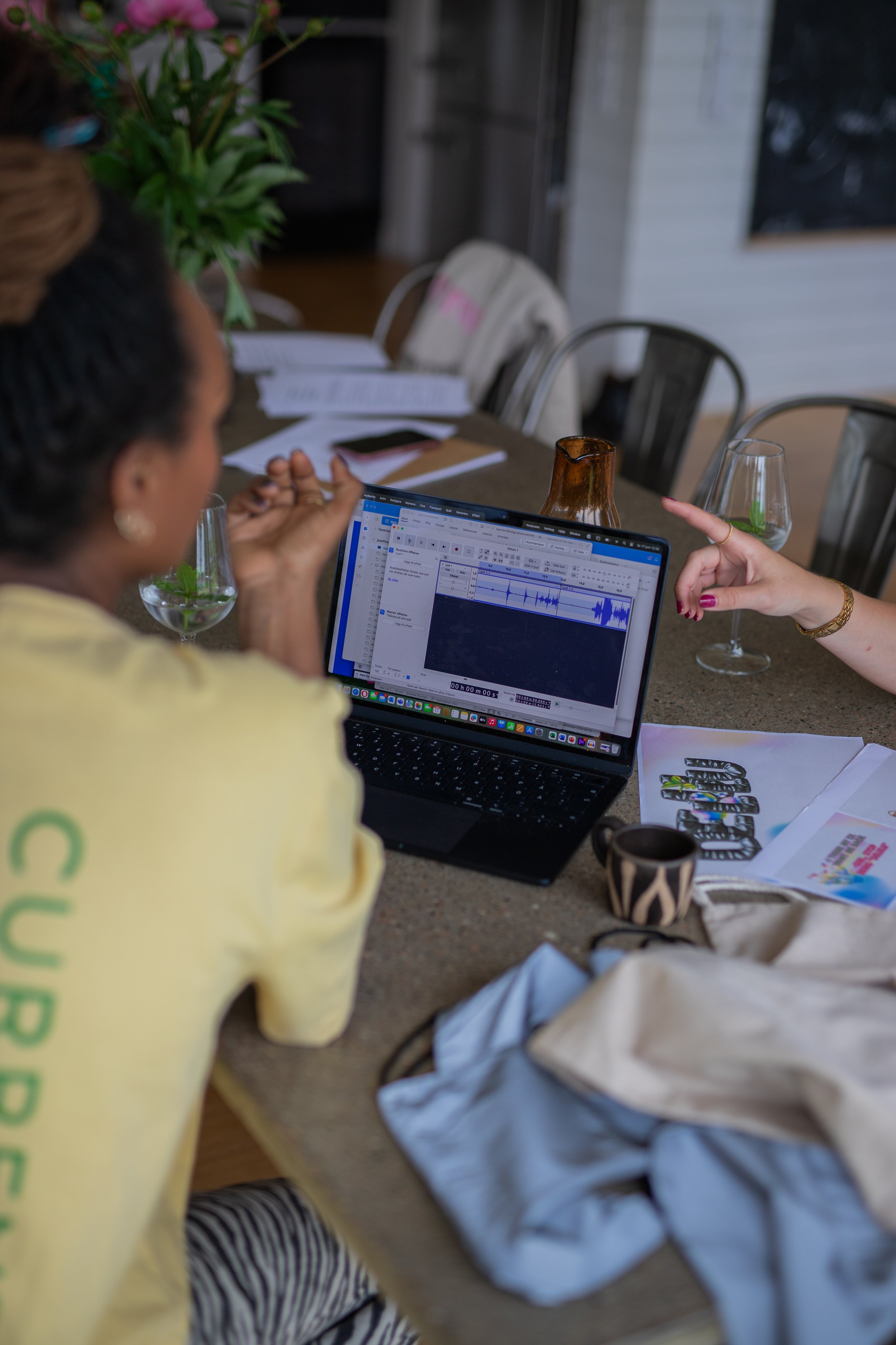 Two women discussing in front of a laptop with audio editing software on a table, surrounded by papers, glasses of water, and other items.