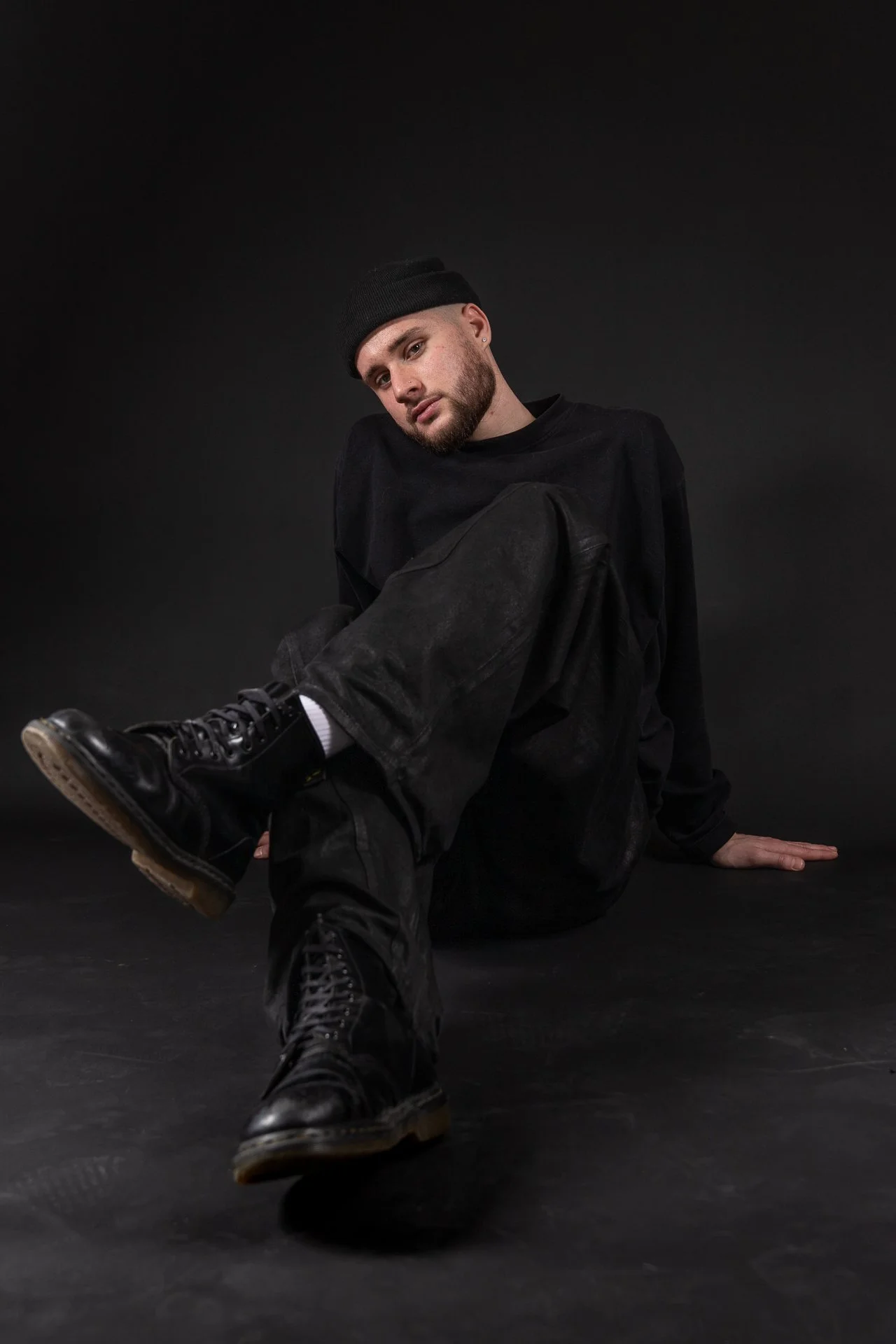 A young man with a beard wearing a black beanie, black sweatshirt, black pants, and black boots, sitting on the floor against a dark background. He is looking at the camera with a relaxed expression.