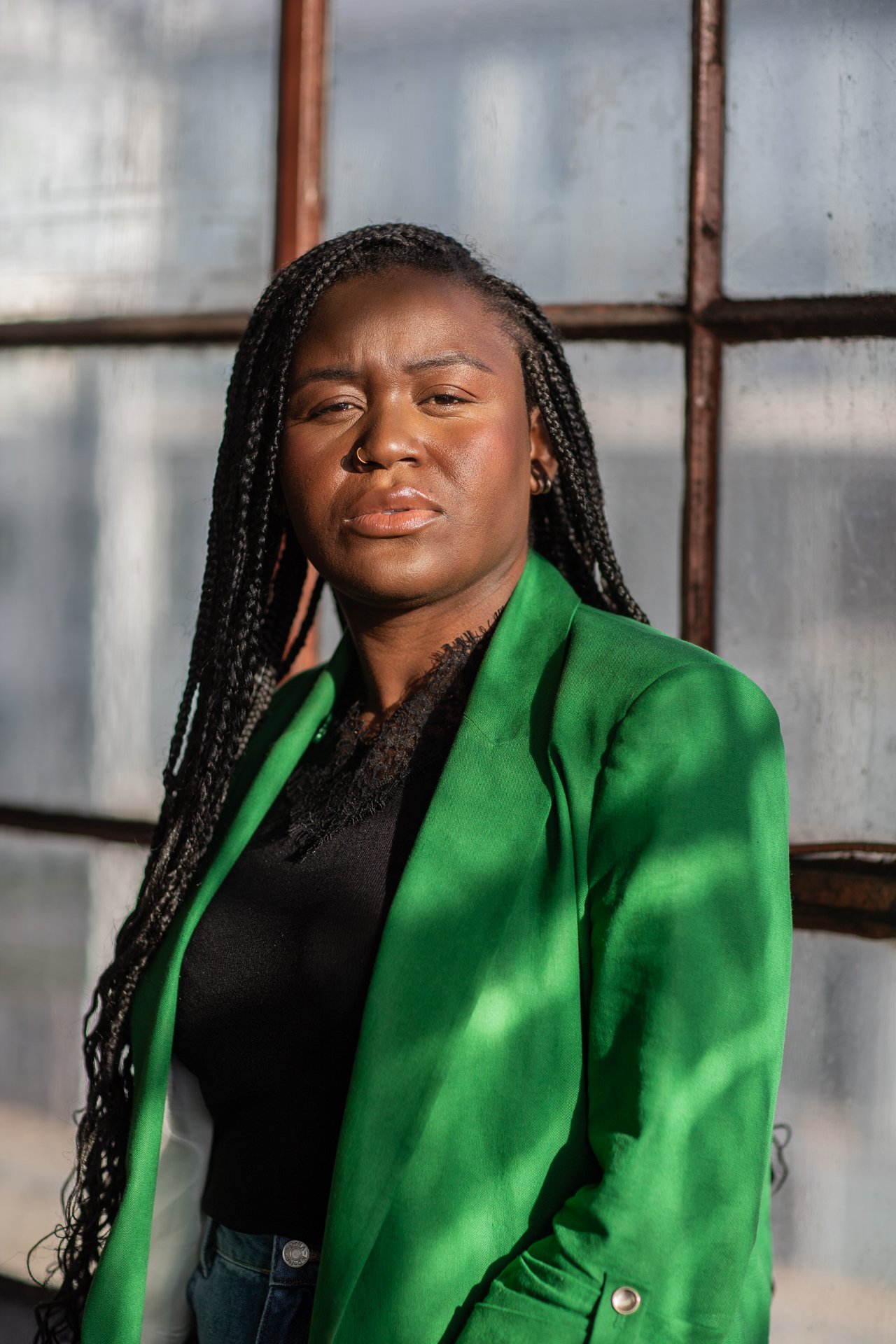 A woman with long braided hair wearing a green blazer over a black top standing in front of a metallic background.