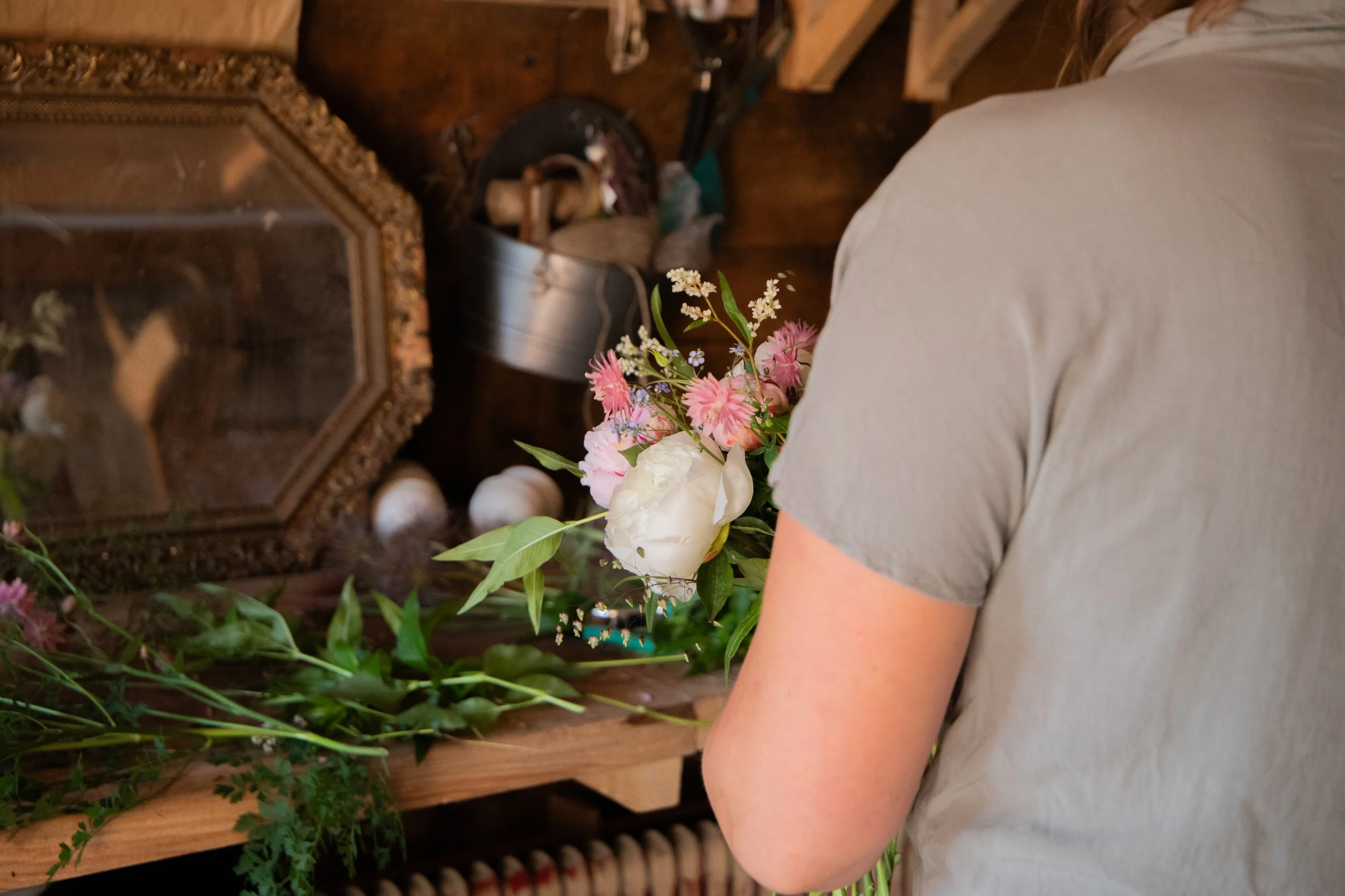 A person holding a bouquet of pink and white flowers with green leaves in front of a wooden shelf with various objects, including a mirror, a metal bucket, and white decorative balls.