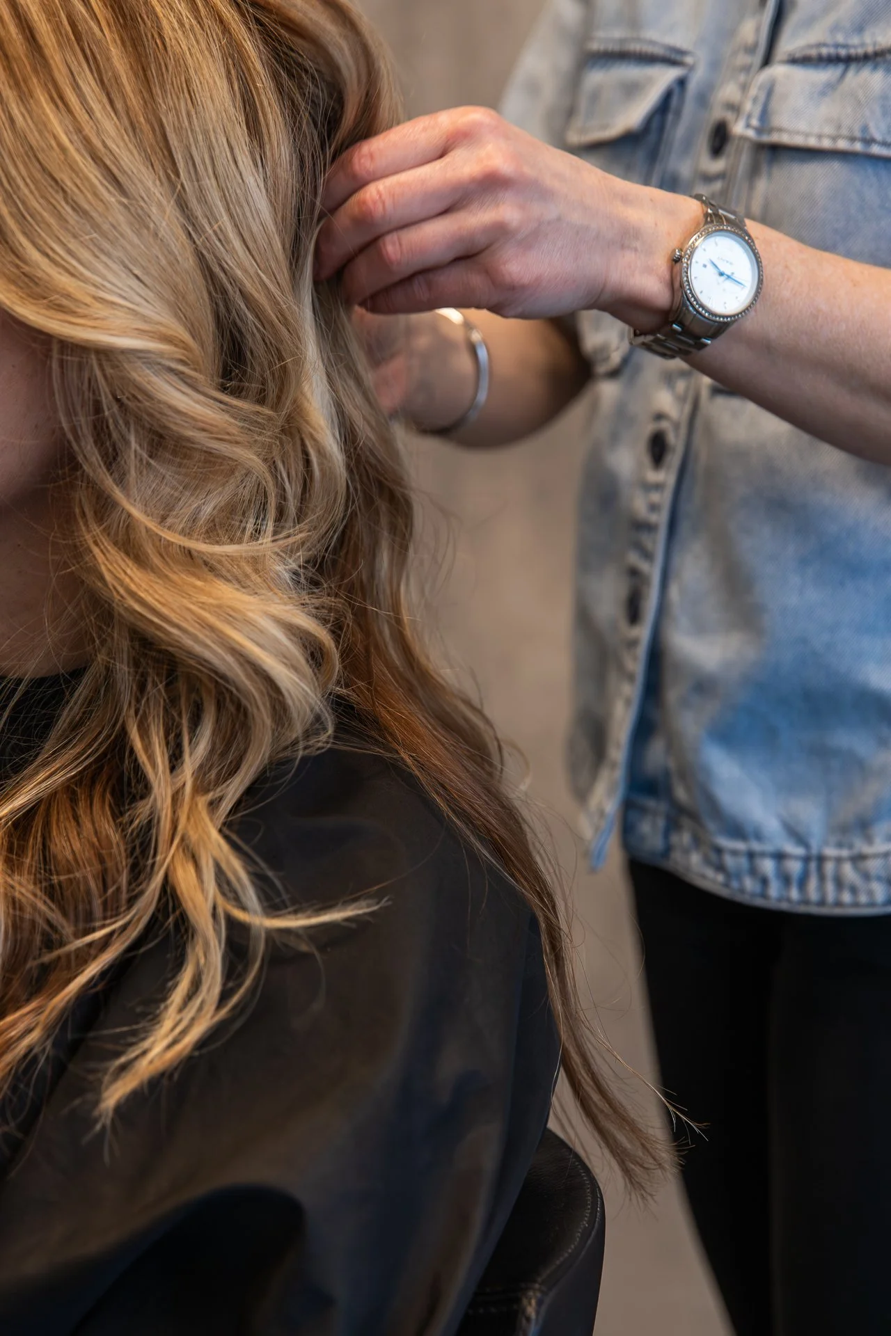 Close-up of a person's hand resting on their head, showing a silver watch on their wrist, next to another person wearing a denim jacket.