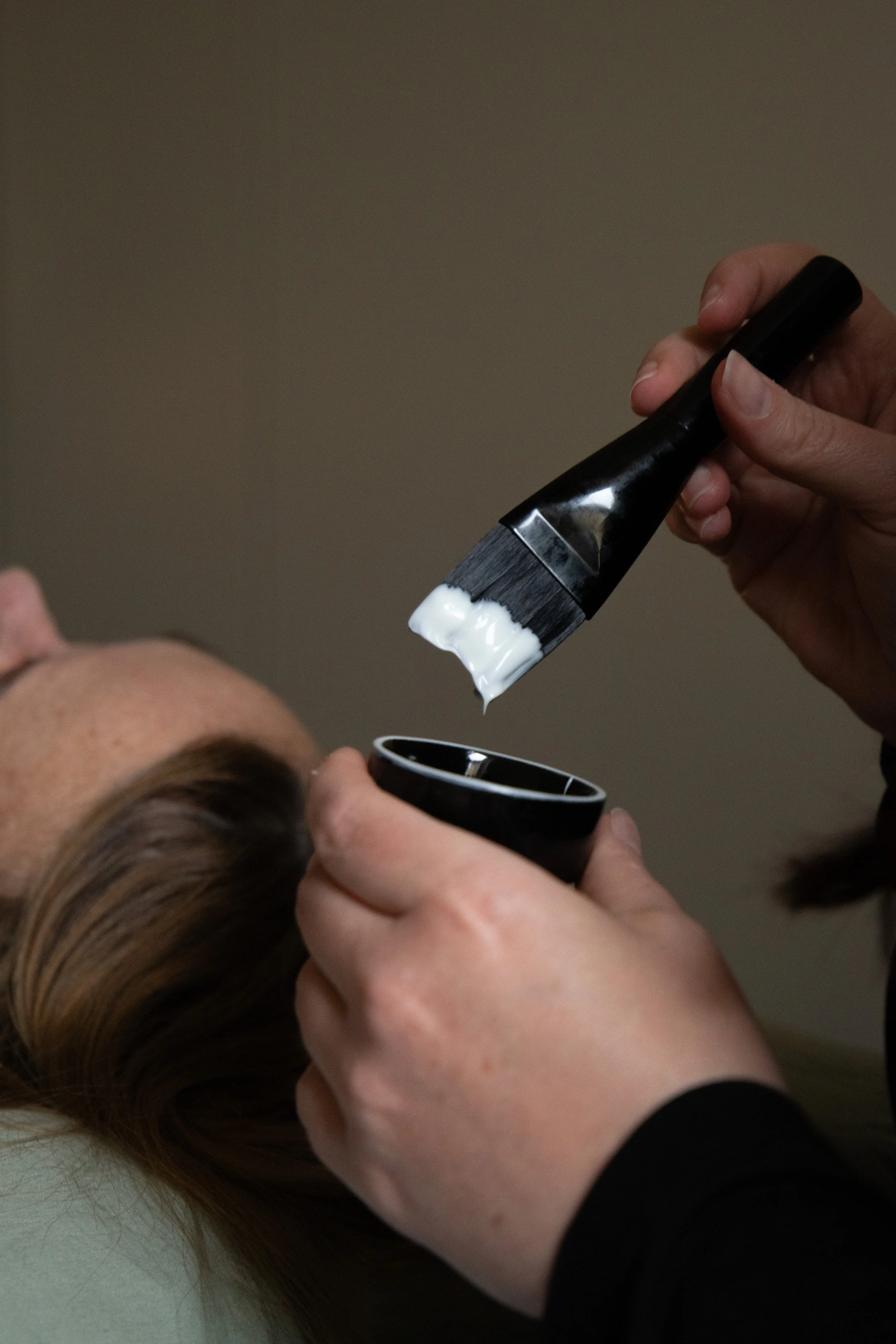 A person applying a white facial or skincare product with a brush to a woman's face, who is lying down.