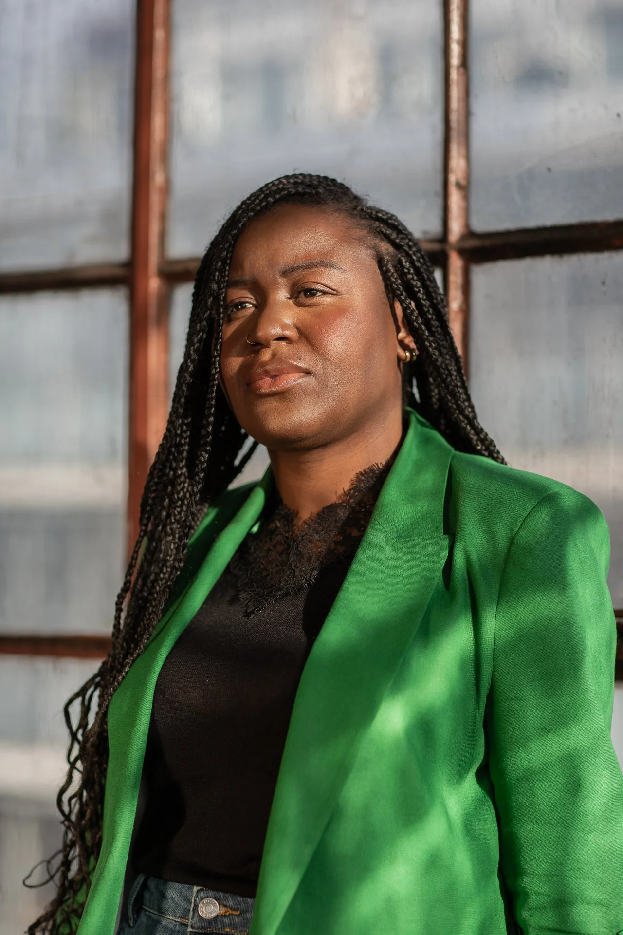 A woman with long braided hair wearing a bright green blazer and a black top, standing indoors near a window with metal framing.