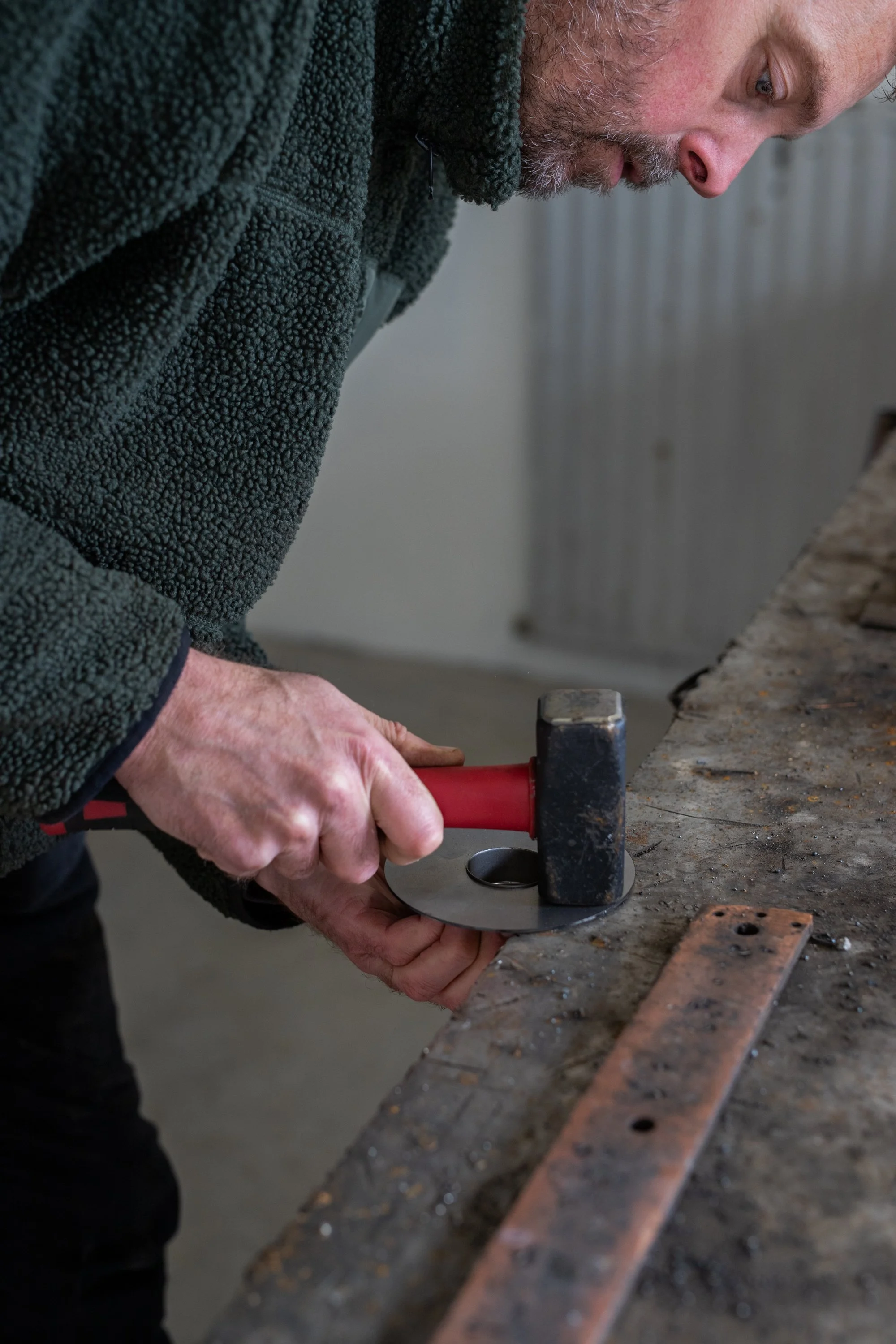 A man wearing a dark fleece jacket is using a hammer and a metal tool on a workbench in a workshop.
