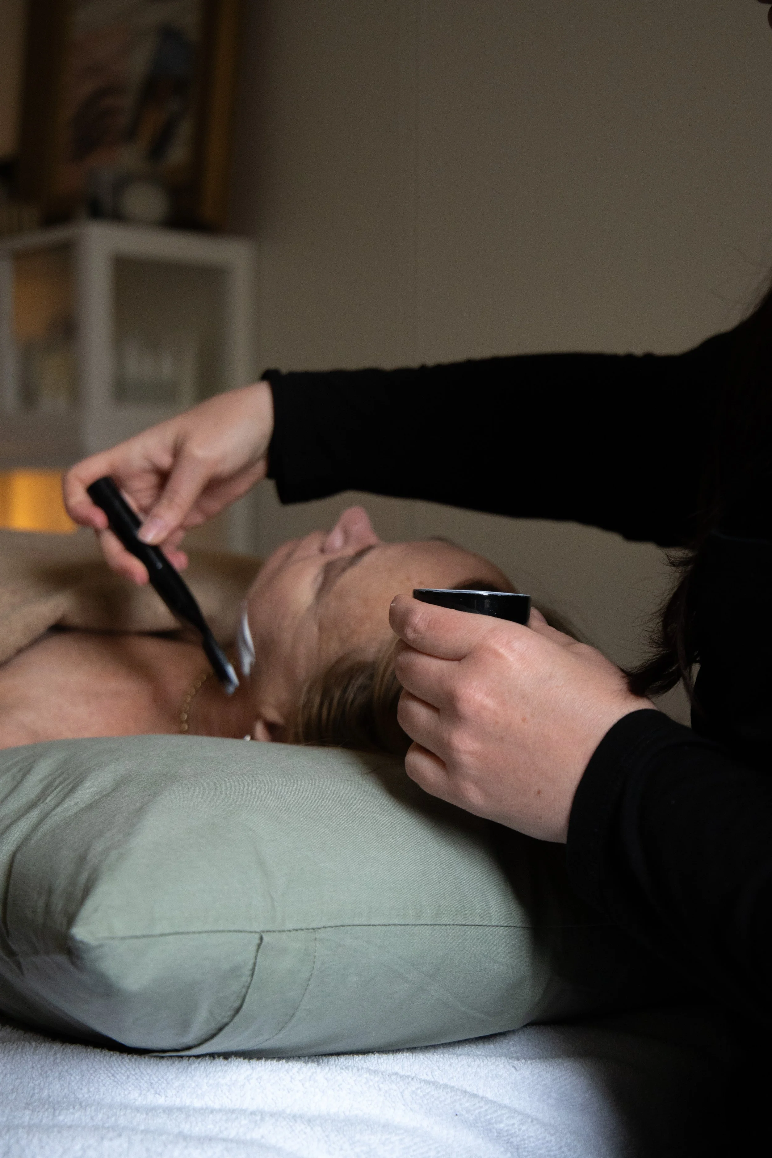 A person lying on a bed with a pillow, receiving makeup application from another person. The makeup artist holds a brush in one hand and a small mirror in the other.