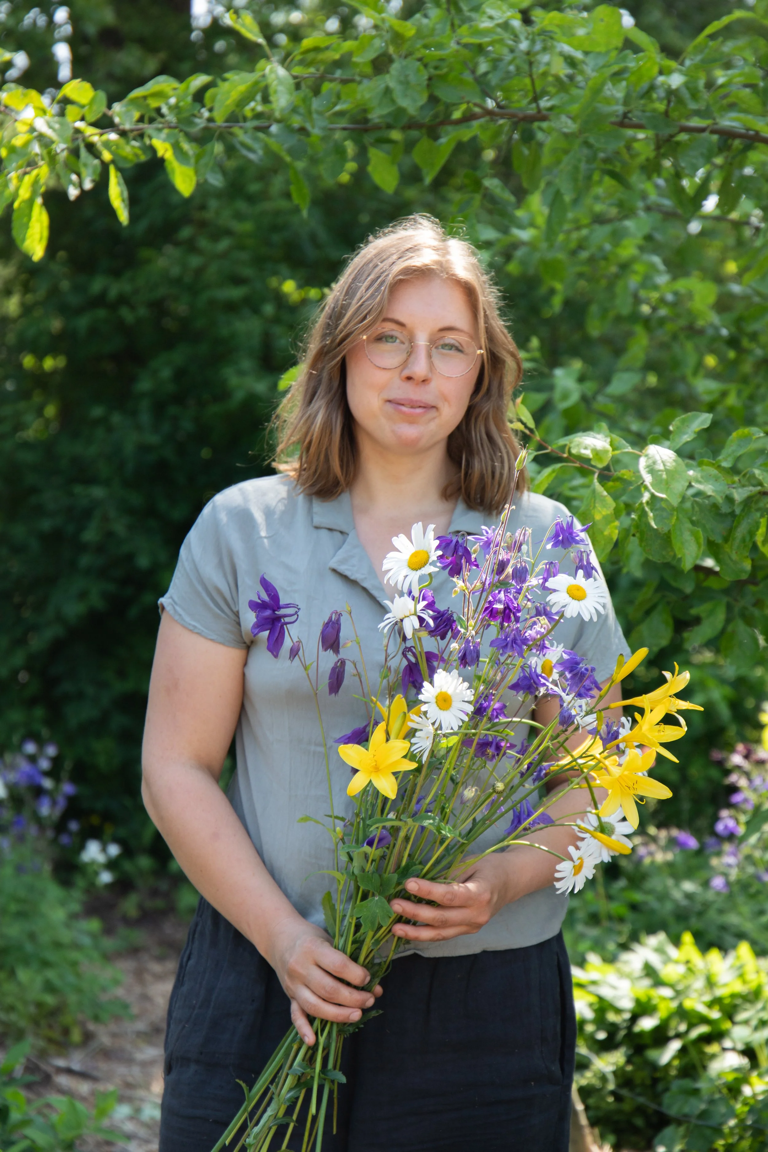 A woman with shoulder-length brown hair, glasses, and a light gray shirt holding a large bouquet of yellow, white, and purple flowers outdoors, standing amidst greenery.