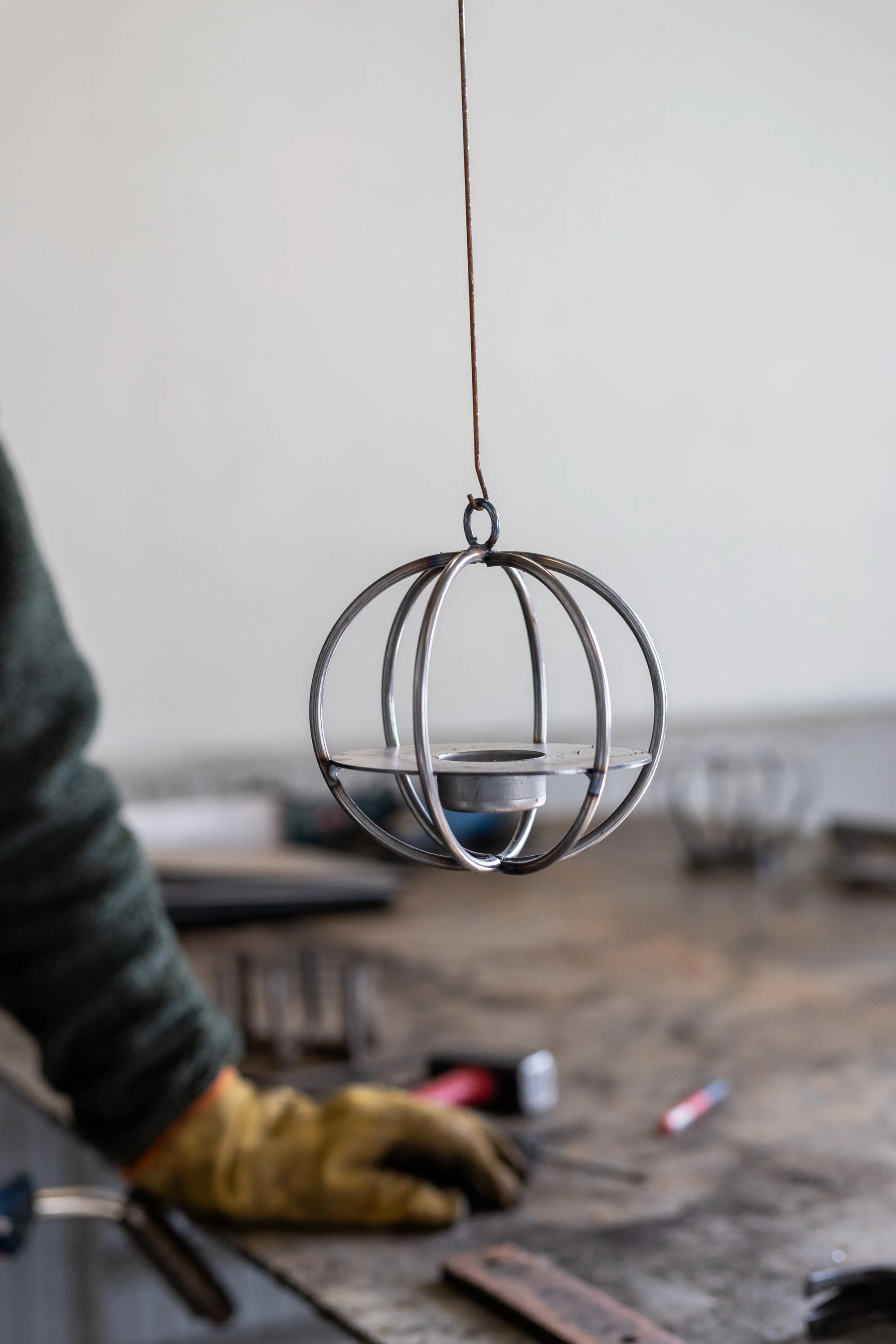 A metal candle holder hanging from a thin wire in a workshop, with a person's gloved hand and work tools visible on a workbench below.