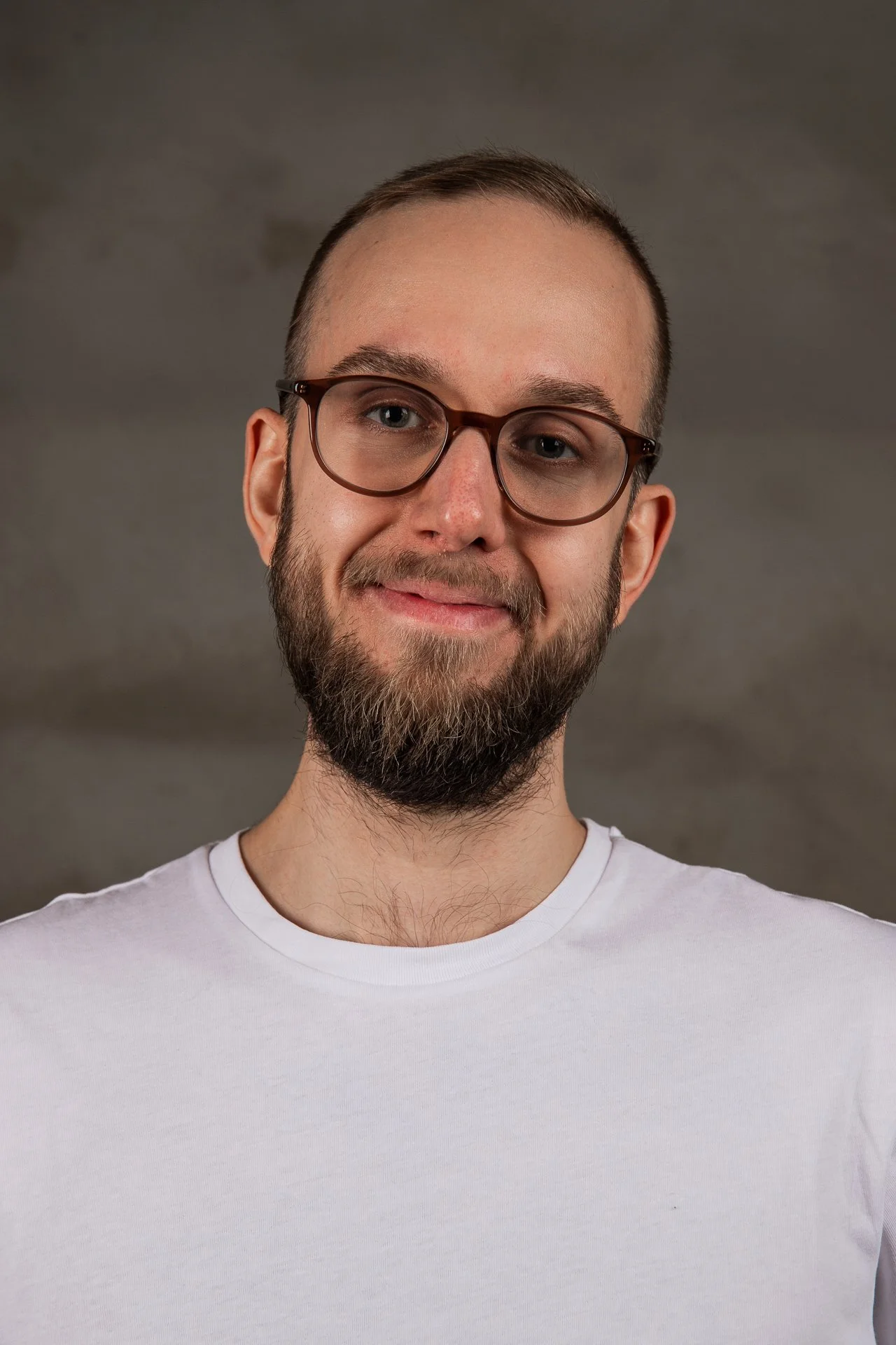 A young man with glasses, a beard, and short hair wearing a white t-shirt, smiling at the camera against a gray background.