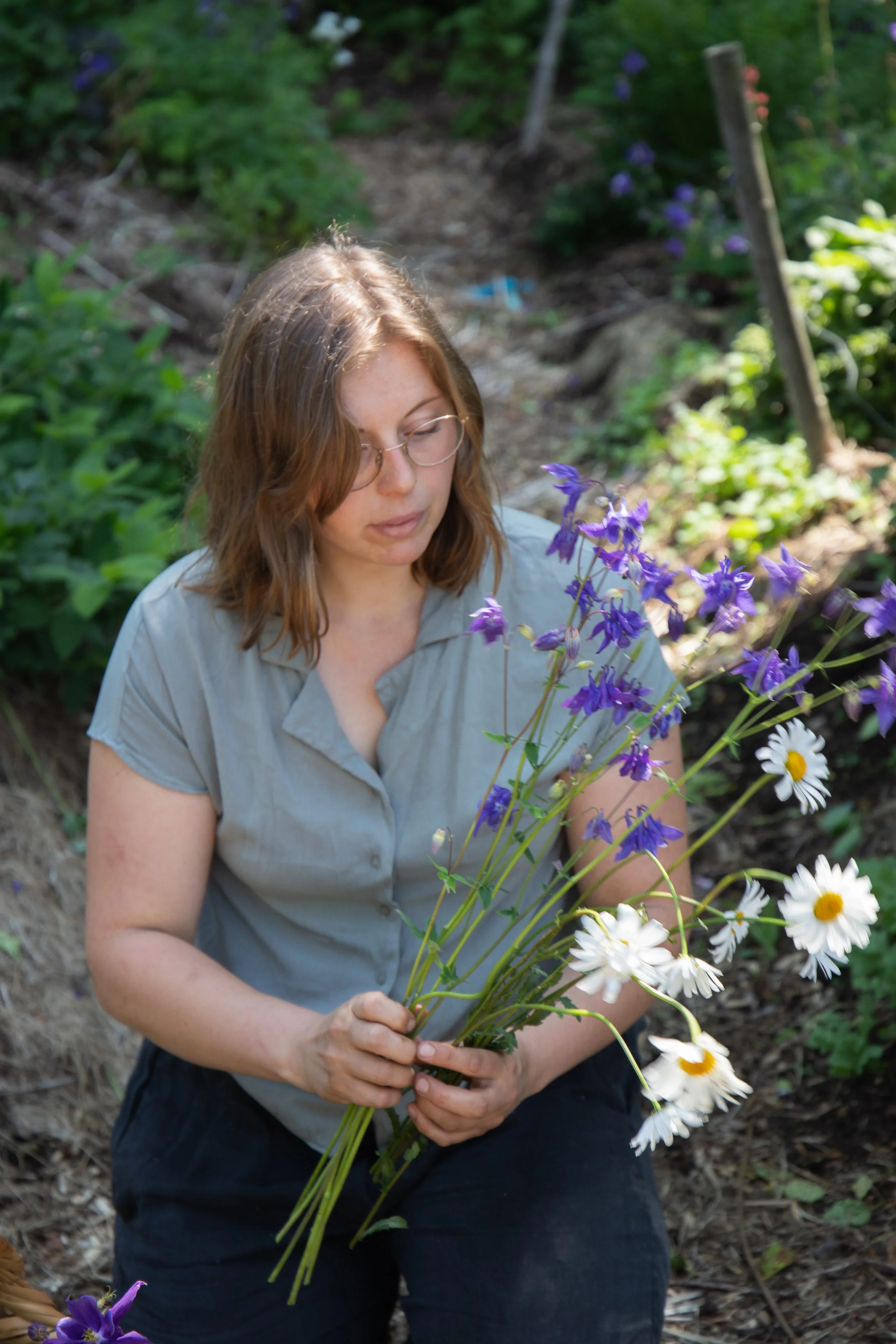 A woman with glasses and shoulder-length brown hair wearing a gray shirt holding a bouquet of purple and white flowers in a garden.