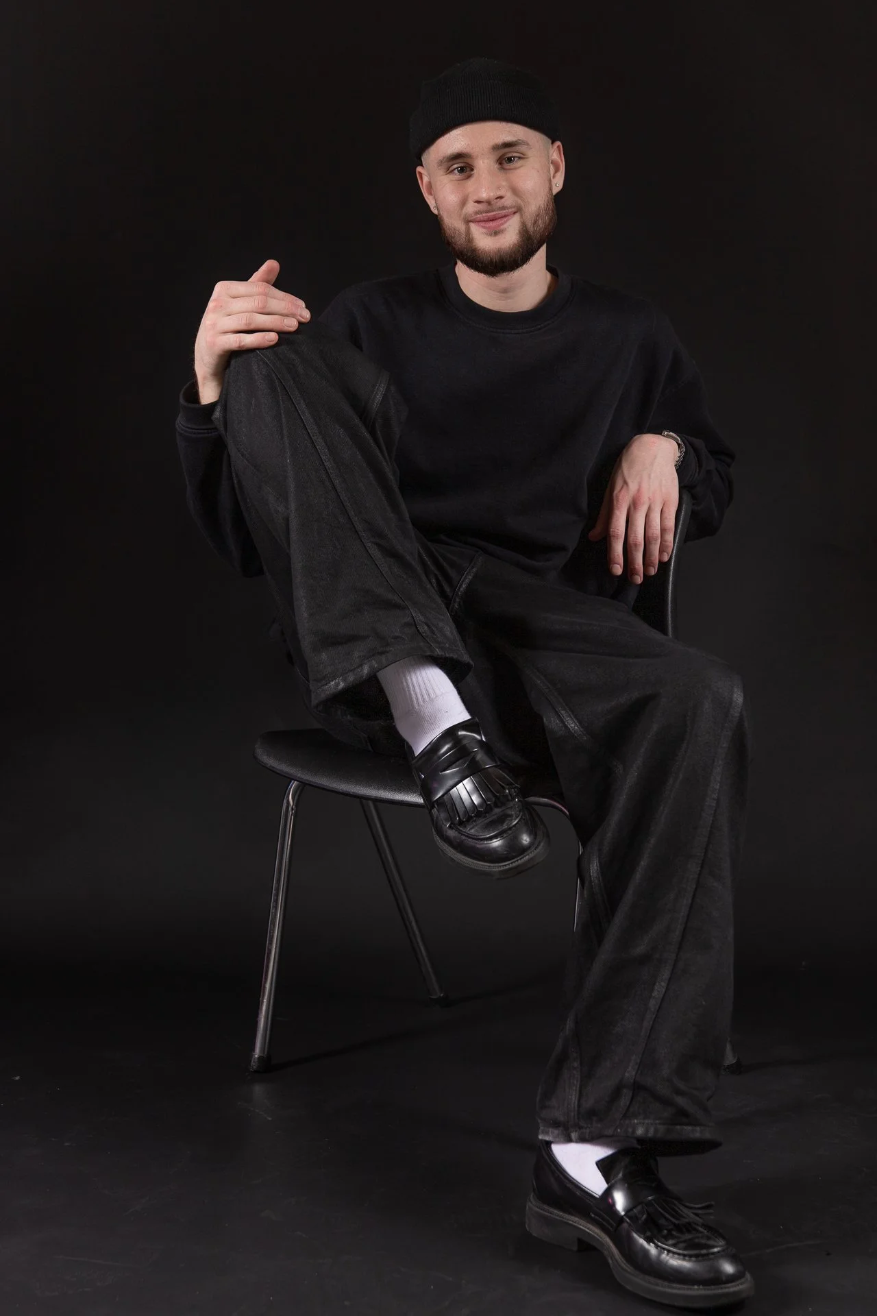 A young man sitting on a black chair against a black background, wearing a black beanie, black sweatshirt, black pants, white socks, and black shoes, with a slight smile on his face.
