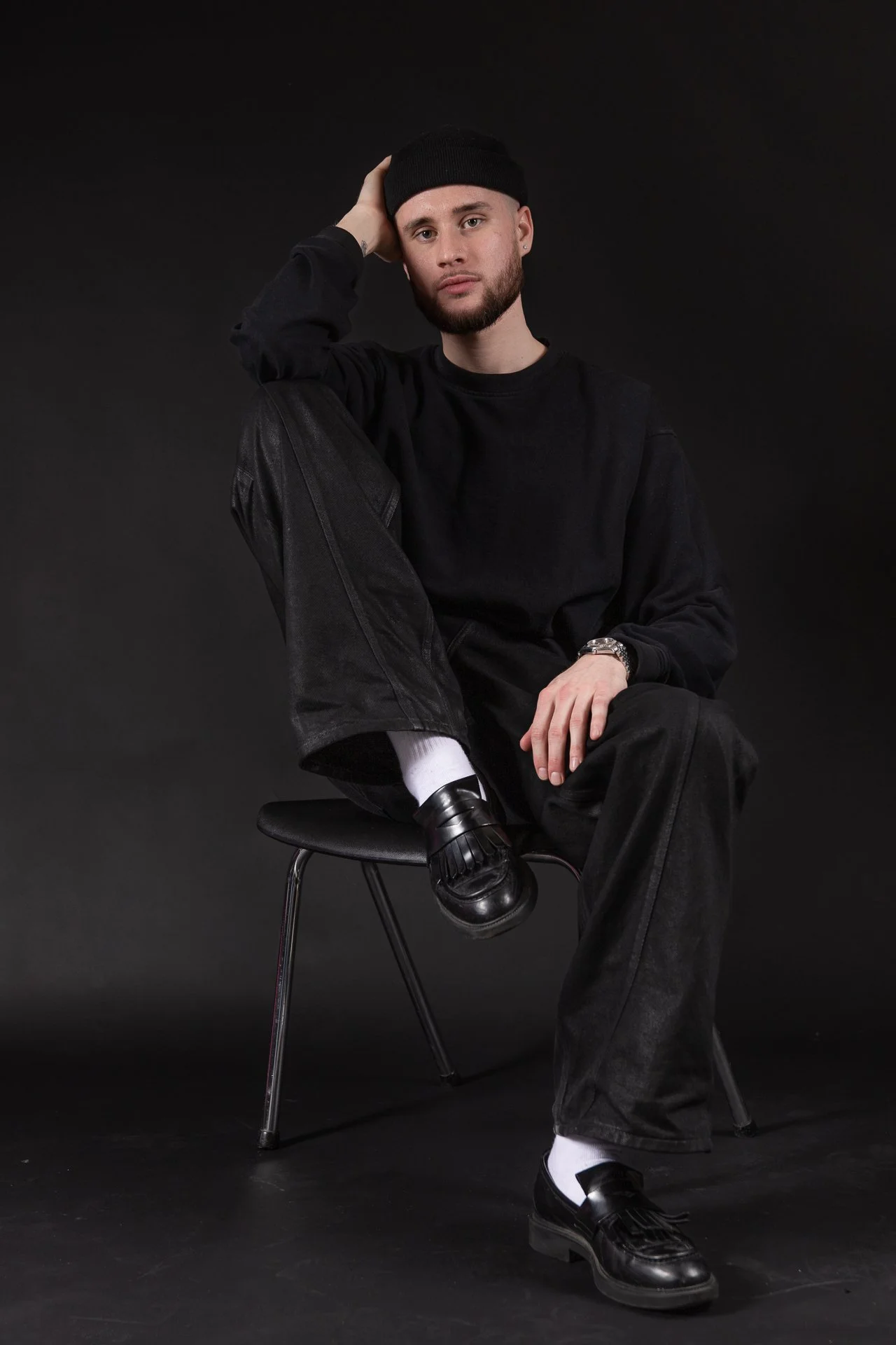 A young man wearing a black beanie, black sweatshirt, black jeans, white socks, and black loafers sitting on a black chair against a dark background. He has a beard and is leaning on his left hand with a relaxed expression.