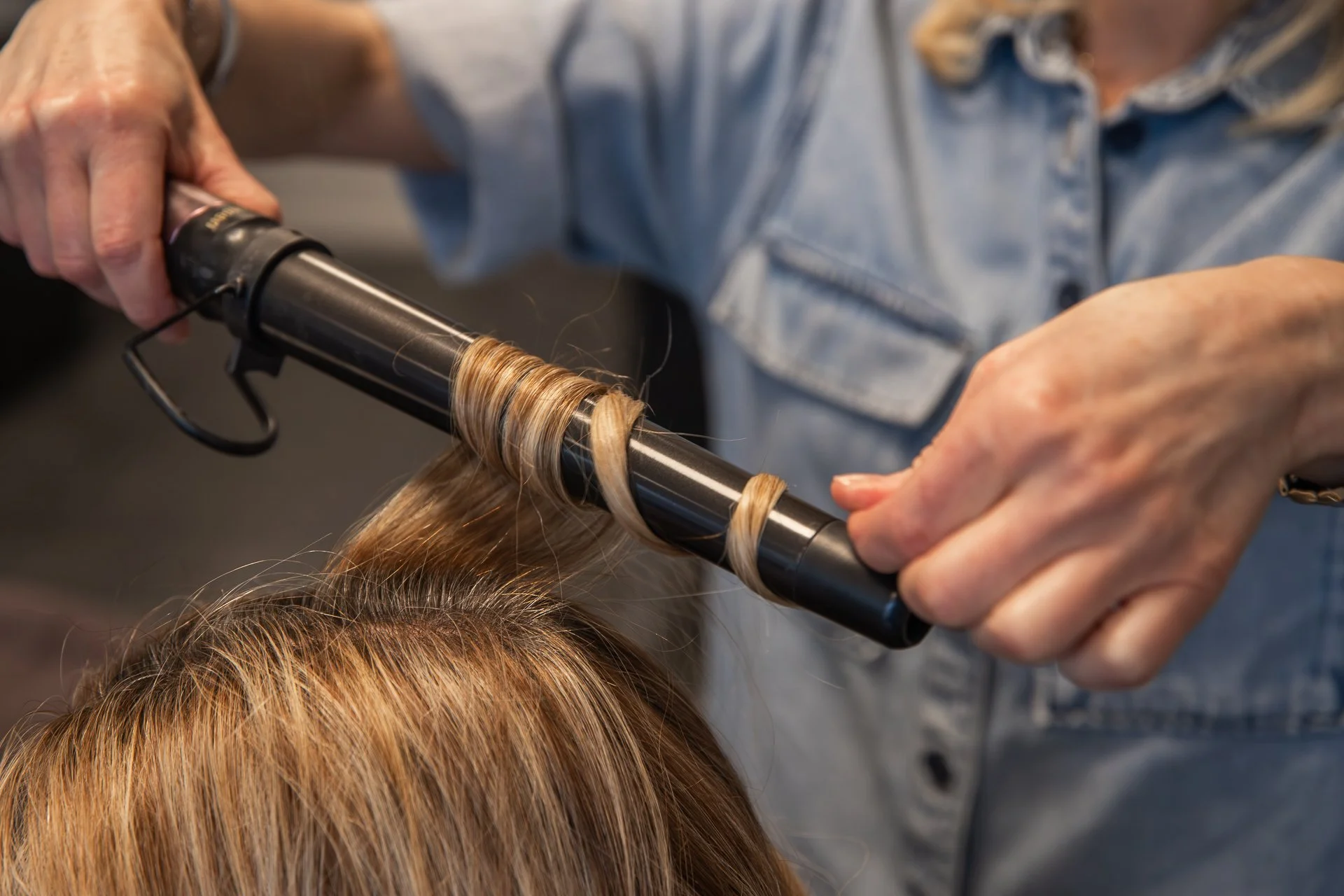 Person styling hair with curling iron, wrapped around a black rod, while wearing a striped shirt.