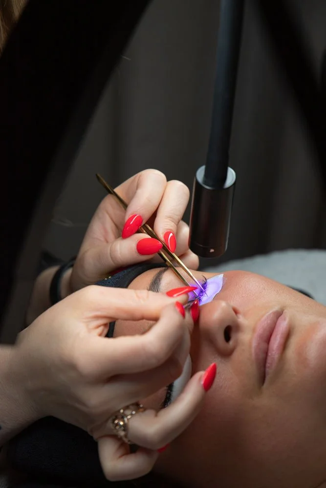 A person receiving a laser eye treatment or eyelash procedure, with a technician using a tool emitting a violet light on the person's closed eyelid.