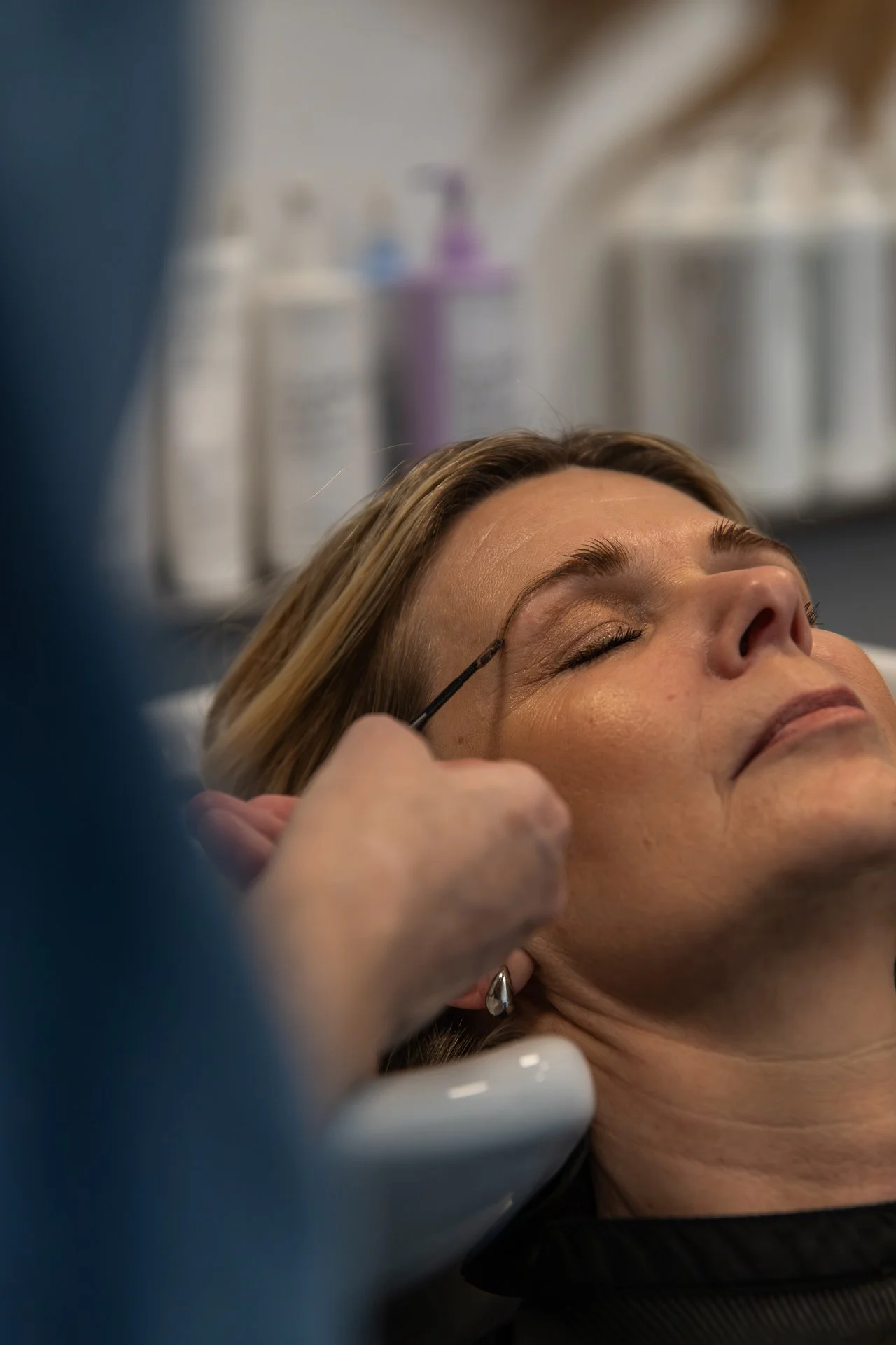 A woman lying with her eyes closed during a cosmetic procedure, with a professional applying makeup or pigment to her eyebrows in a clinical setting.