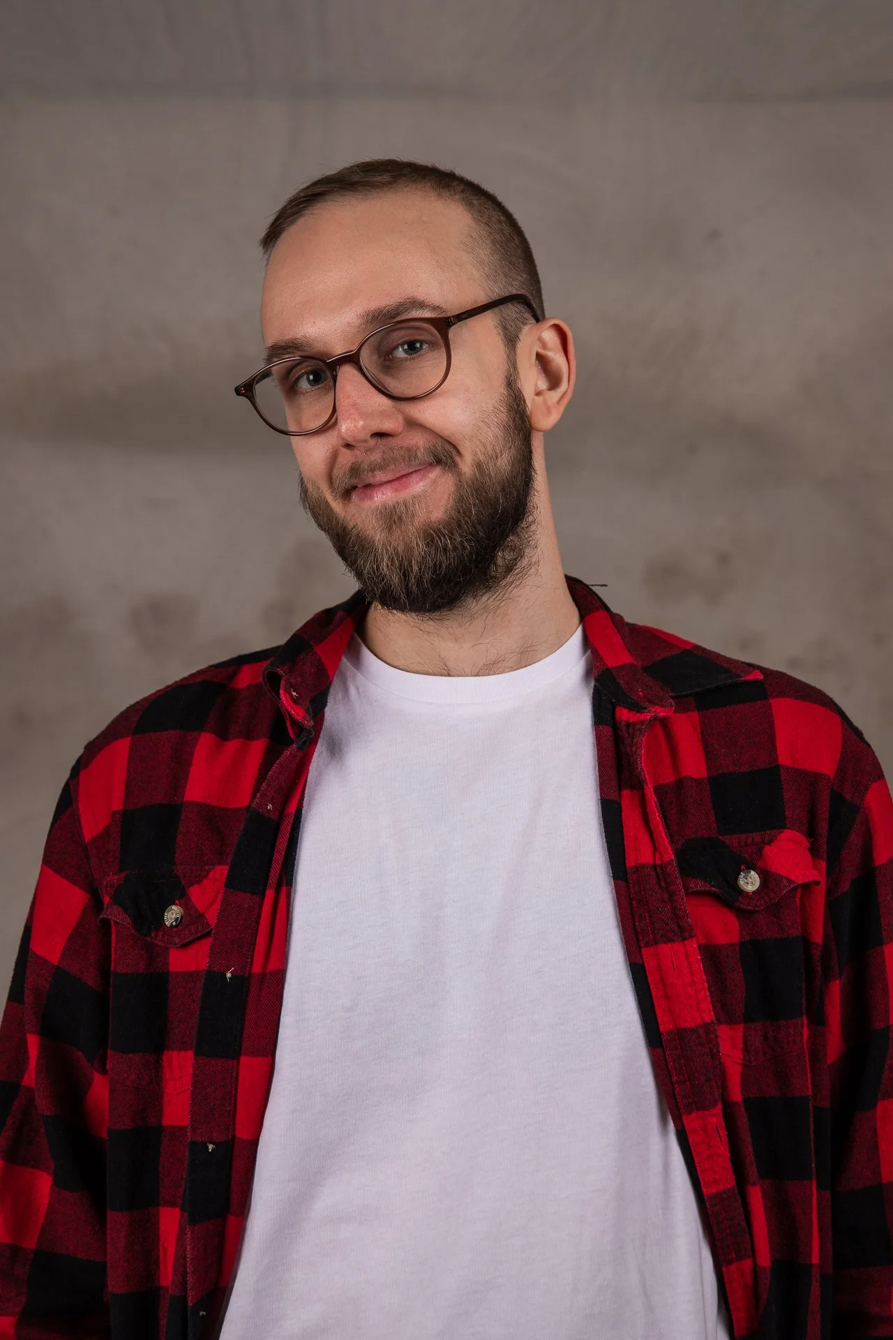 A young man with glasses and a beard, wearing a red and black checkered shirt over a white t-shirt, smiling at the camera against a plain background.