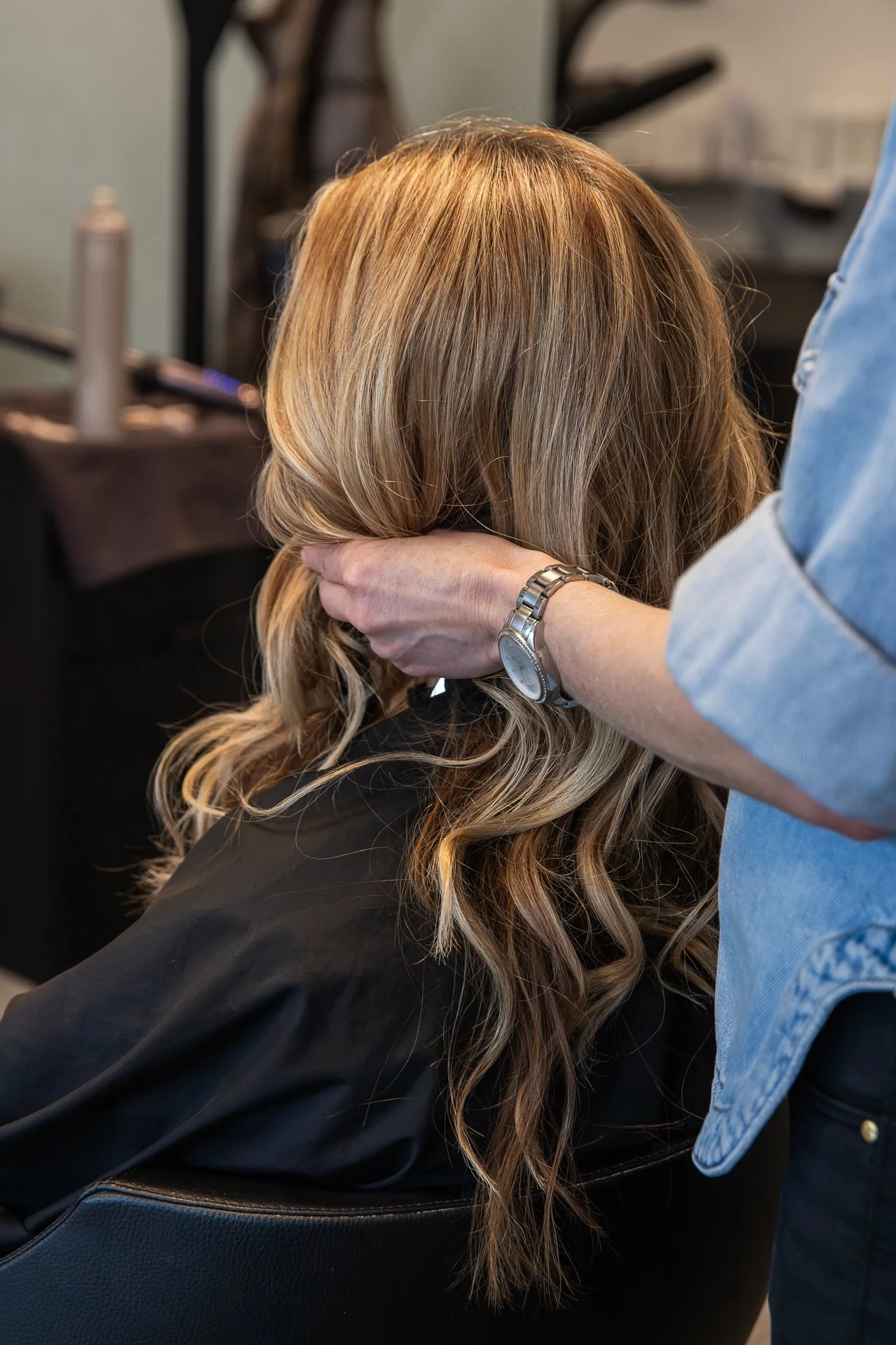 A hairstylist with a silver watch is gently touching a woman's blonde, wavy hair at a salon, with styling tools and products visible in the blurred background.