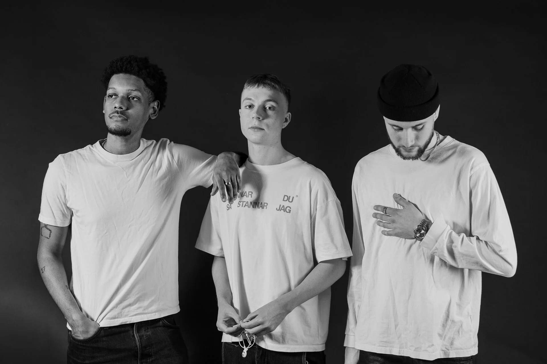 Black and white photo of three young men standing against a dark background. The man on the left has short curly hair and is wearing a white T-shirt. The man in the middle has short straight hair and is wearing a T-shirt with text on it. The man on t
