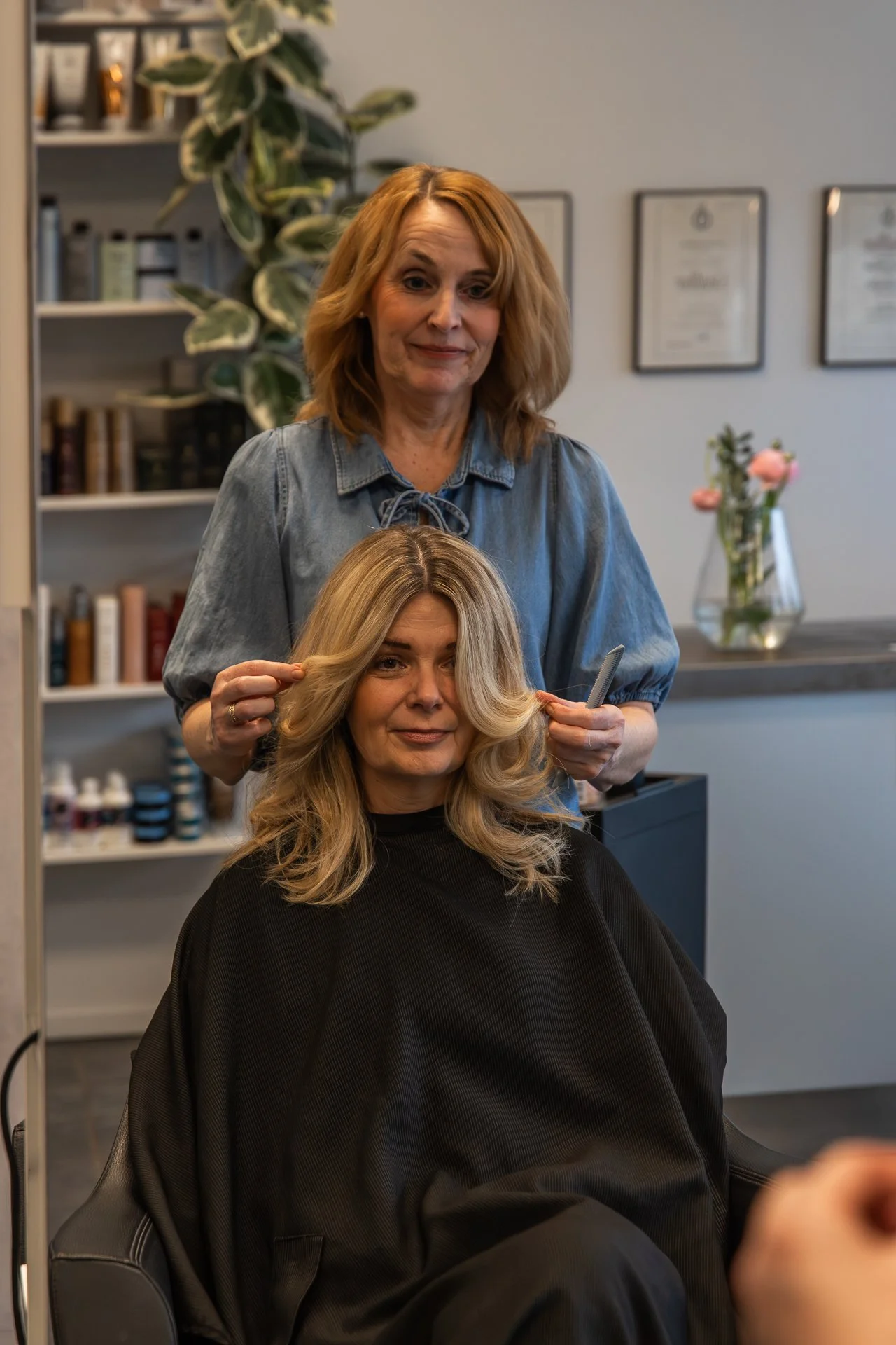 A hairdresser is styling a woman's long, blonde hair while she sits in a salon chair. The woman has a black cape around her and is smiling slightly. The hairdresser is standing behind her, holding a section of her hair and a comb. The background incl