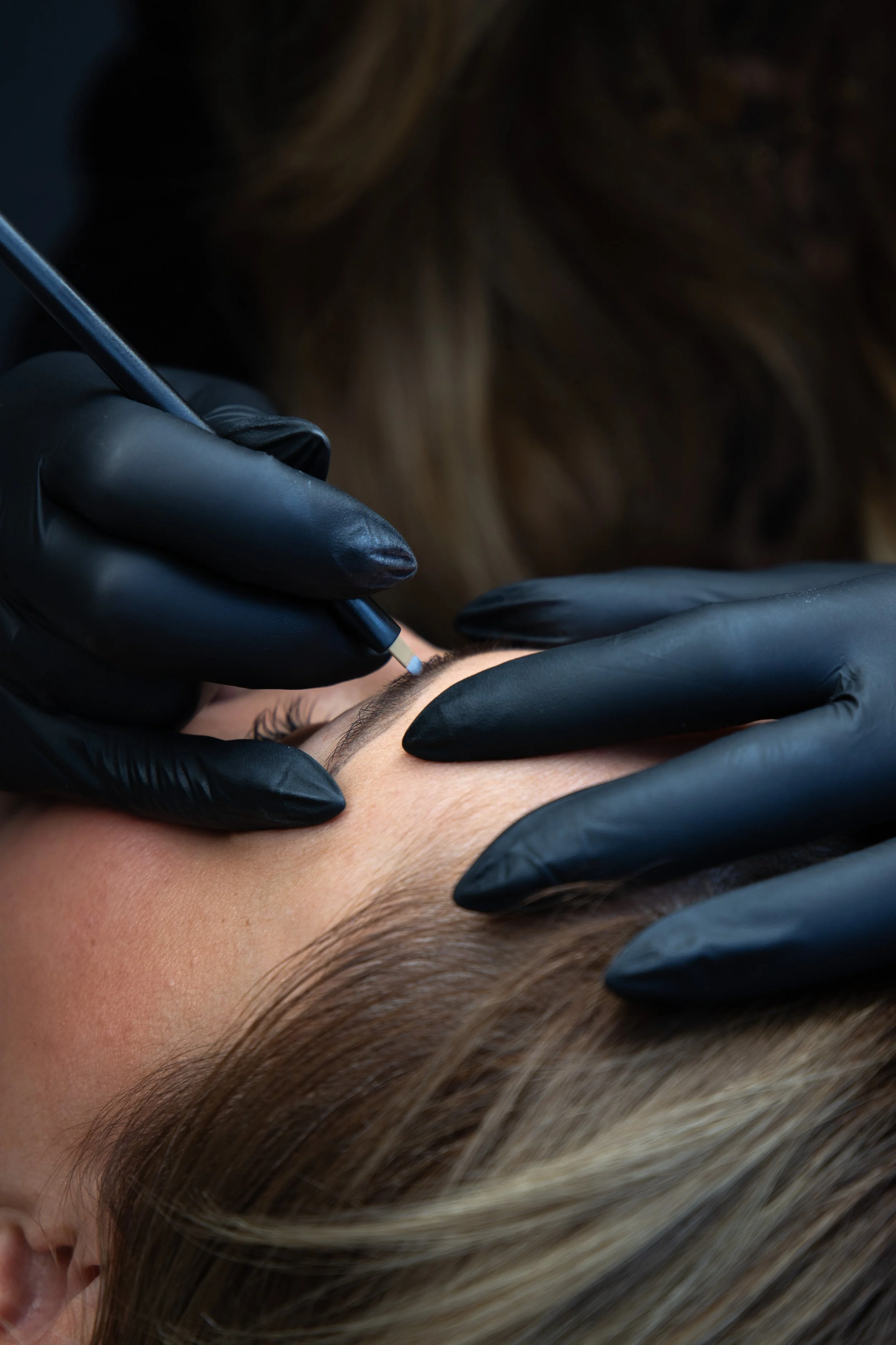 A person receiving a cosmetic tattoo or permanent makeup procedure on their eyebrows from an artist wearing black gloves.