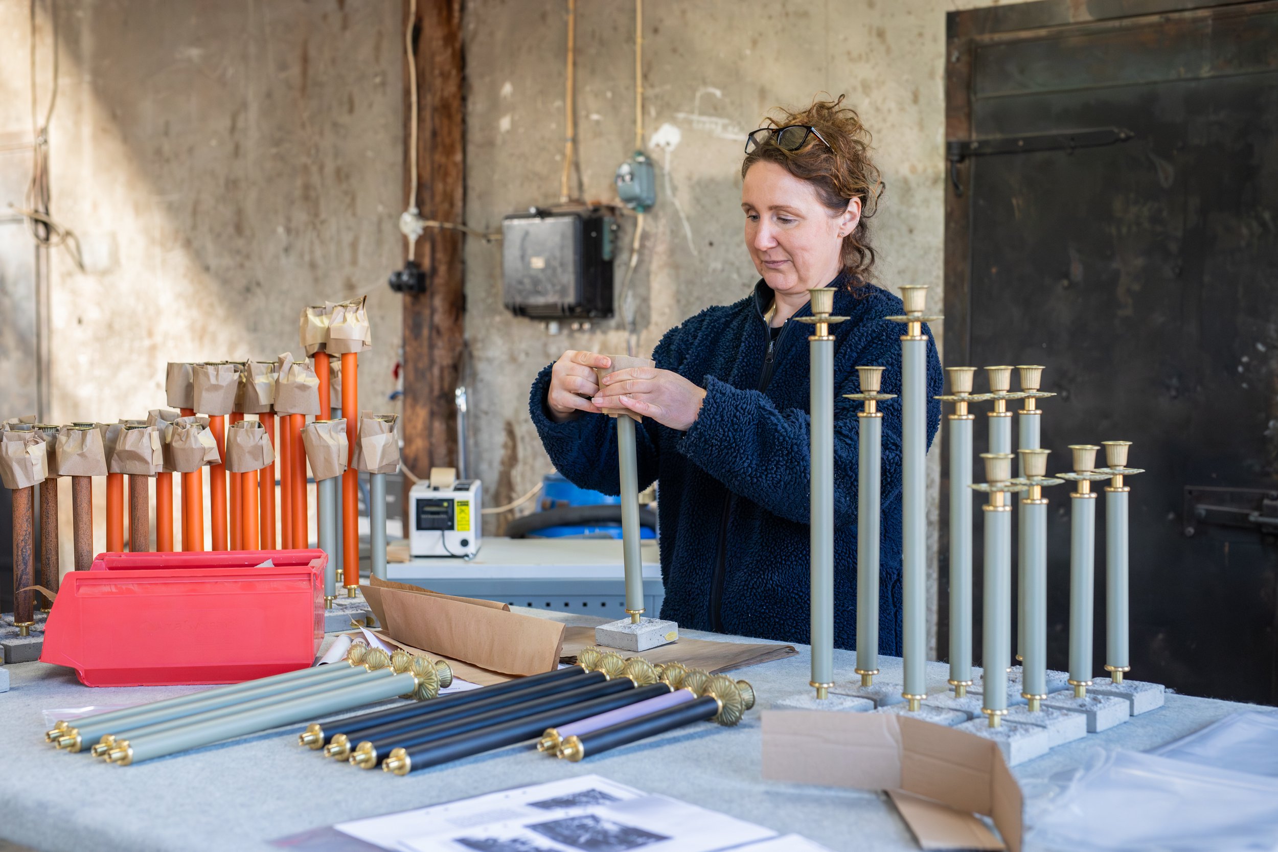 A woman working on assembling or inspecting metal candle holders or similar objects in a workshop with various metal parts and tools on the table.