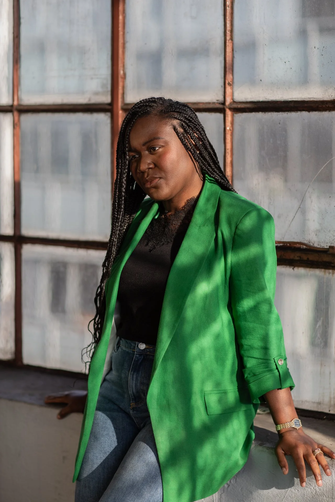 A woman with long braided hair wearing a green blazer and black top, leaning against a metal and glass wall.