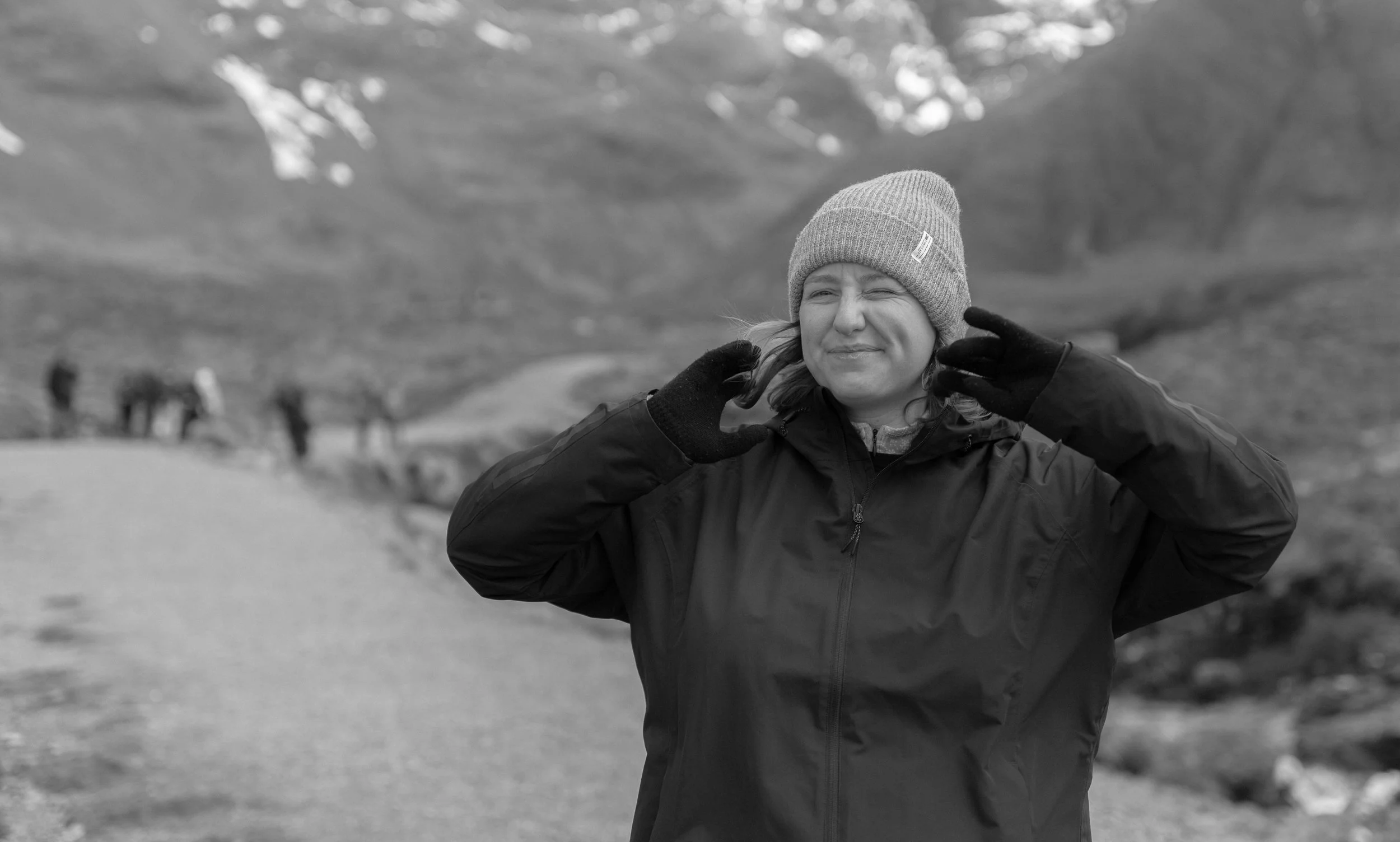 A woman wearing a beanie, gloves, and a jacket making a playful face, squinting her eyes and pushing her cheeks up, outdoors with a blurred mountain landscape and other people in the background.