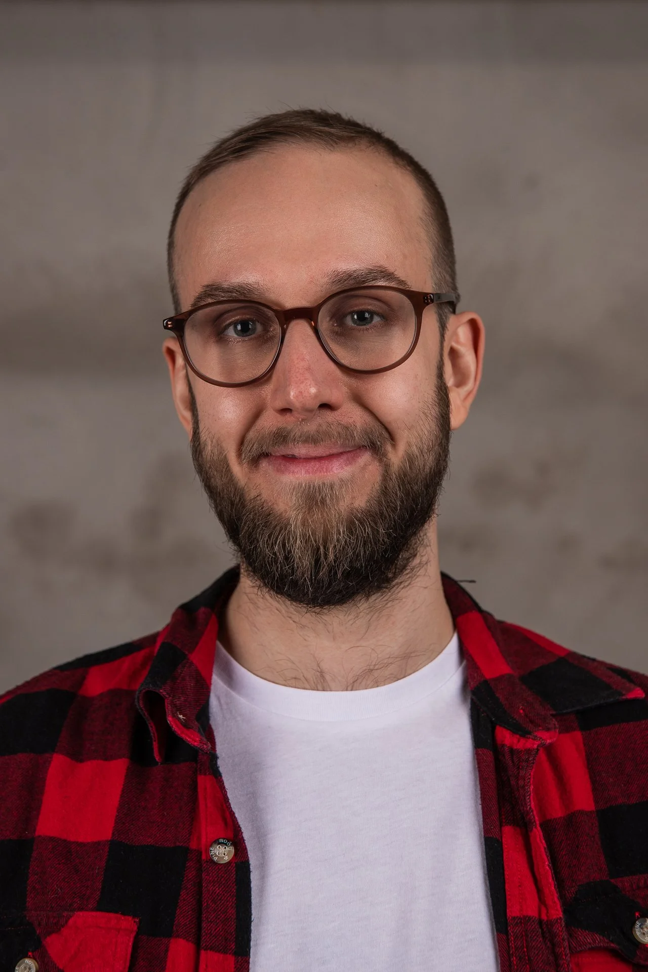 A young man with glasses, a beard, and short hair, smiling and wearing a red and black checkered shirt over a white t-shirt, standing against a plain background.