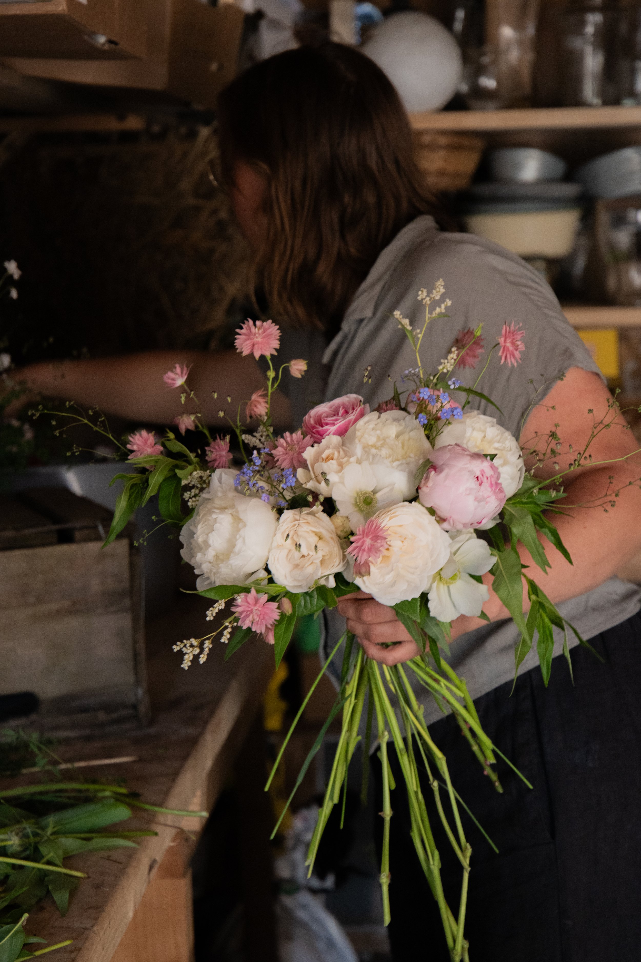 A person holding a bouquet of pink and white flowers, working in a rustic space with wooden shelves and various household items in the background.