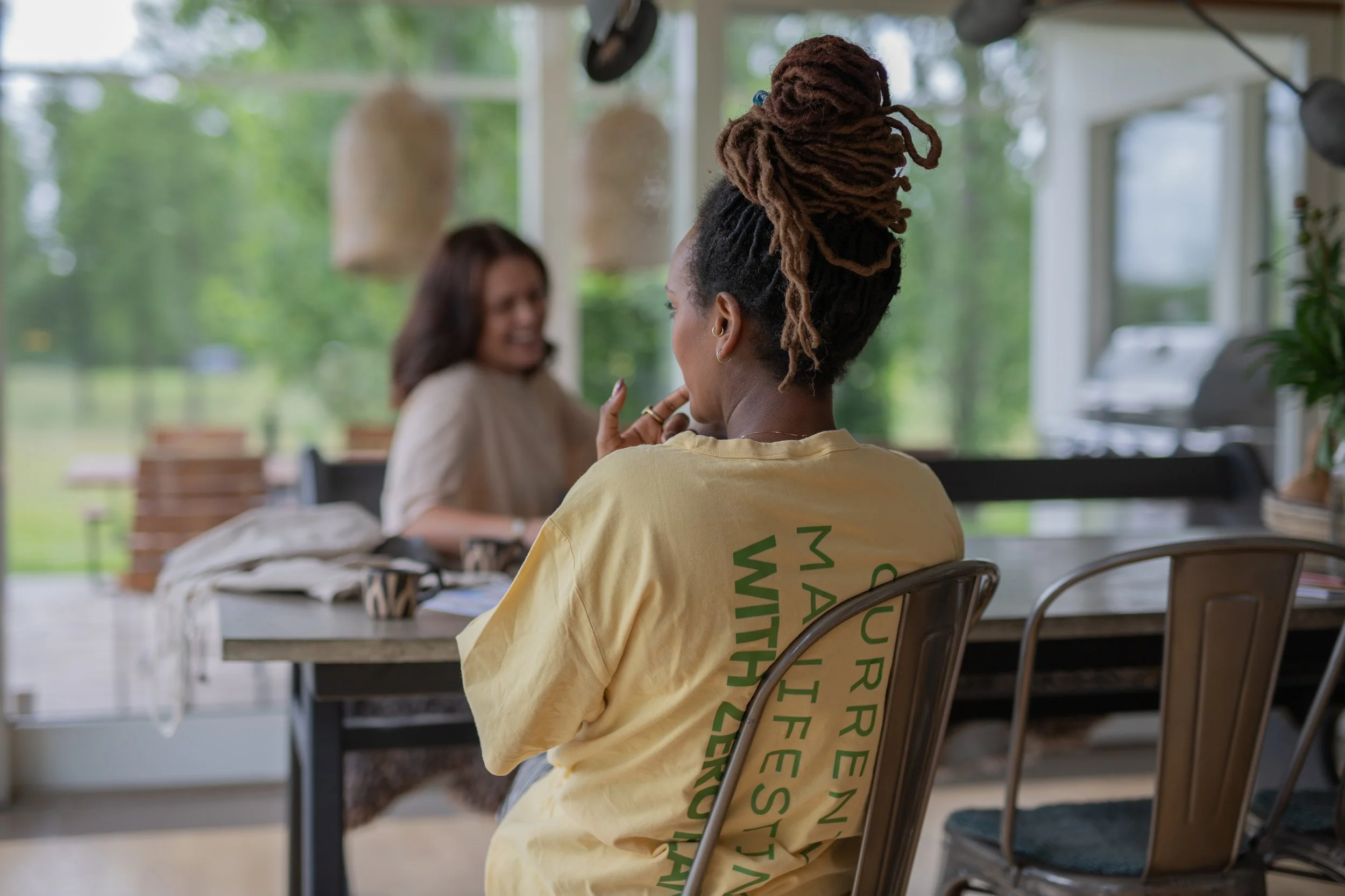Two women sitting at a dining table indoors, with a view of greenery outside through large windows. One woman, with braided hair in a bun, is facing away from the camera wearing a yellow t-shirt with green text. The other woman, with shoulder-length 