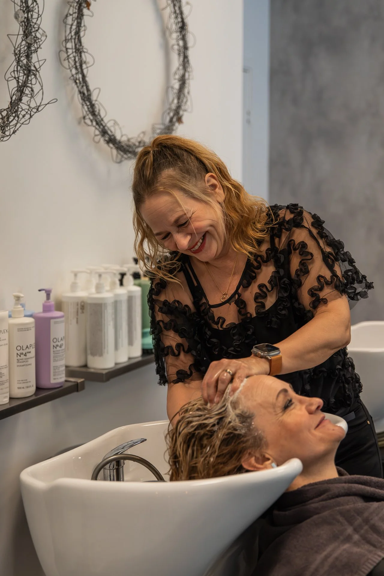A woman with curly red hair is smiling while washing a client's hair at a salon sink. The client is reclined in the sink with her eyes closed. The salon has shelves with various hair care products in bottles.