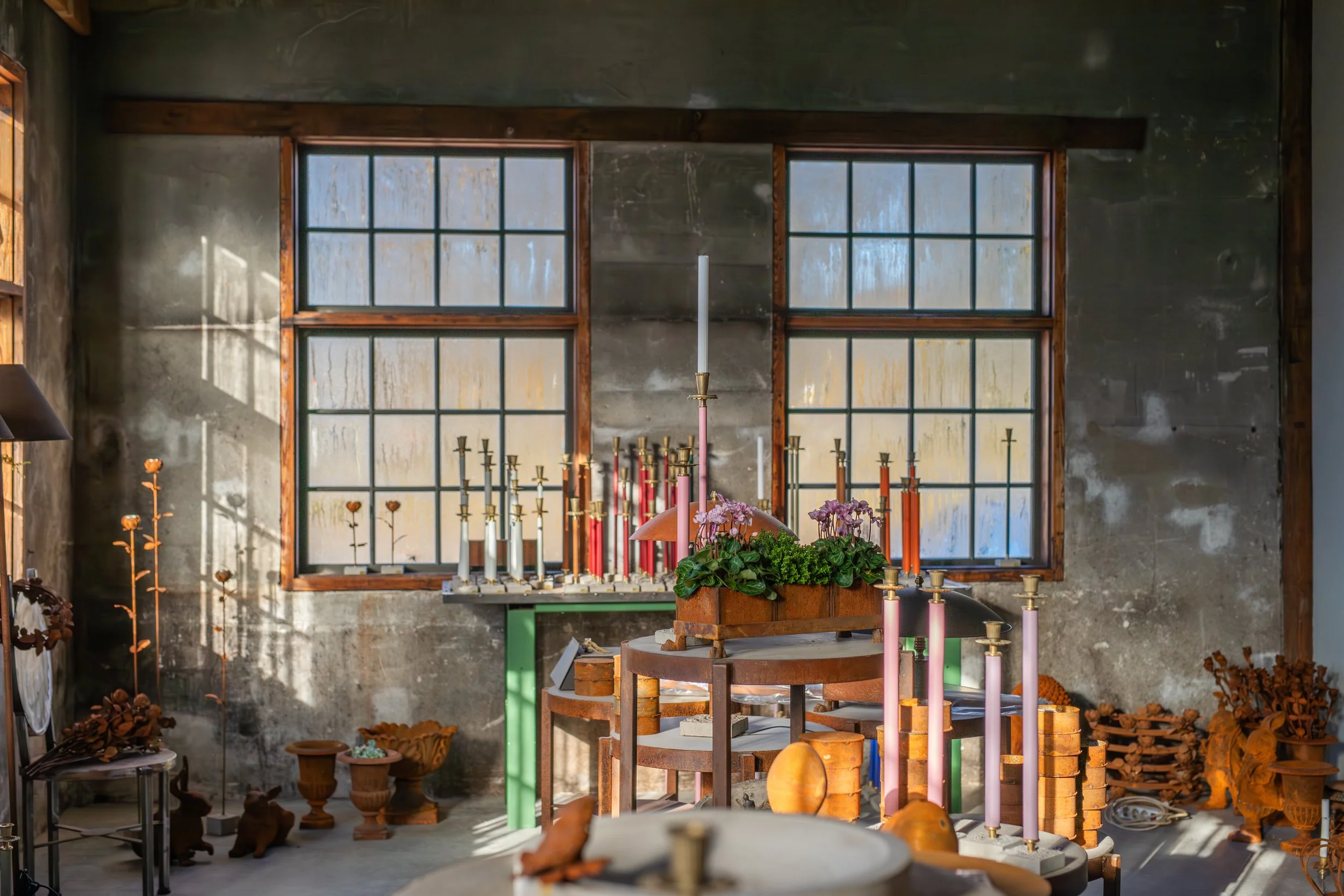 An indoor space with large industrial-style windows, a table with a floral arrangement and pink candles, and various decorative wooden sculptures and plants.